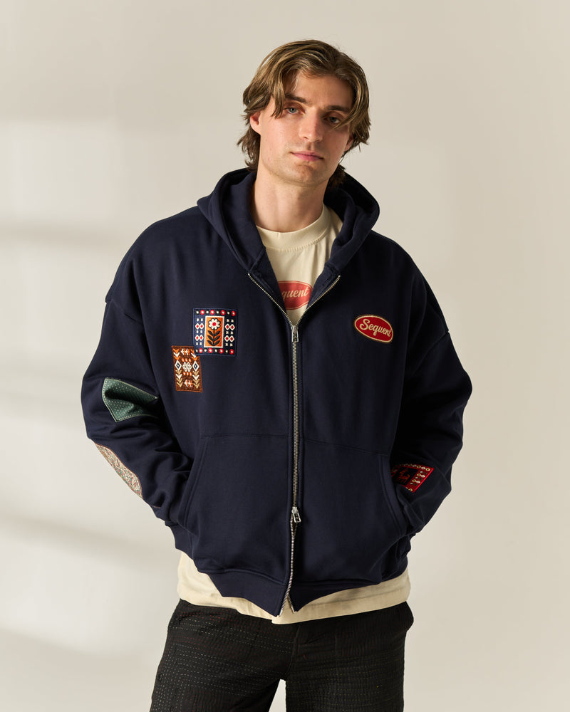 A man wearing a navy blue zip-up hoodie with multiple colorful patches, layered over a cream t-shirt and dark pants, standing against a neutral beige background.