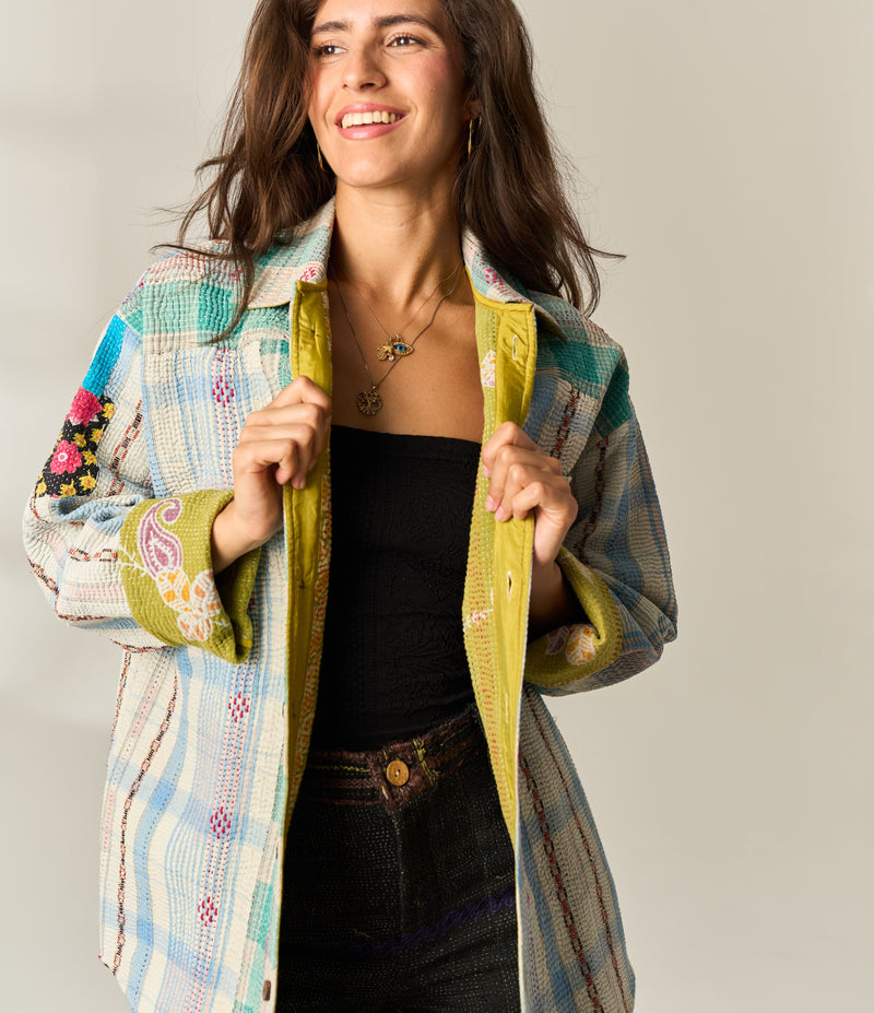 A model viewed from behind wearing a long-sleeve button-up vintage kantha shirt with a colorful, painterly horizontal stripe pattern in orange, blue, pink, and gray, paired with black jeans against a warm off-white background.