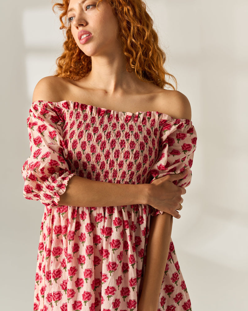 A woman with curly red hair wearing an off-the-shoulder pink hand block printed dress with a red rose pattern, photographed against a neutral light background.