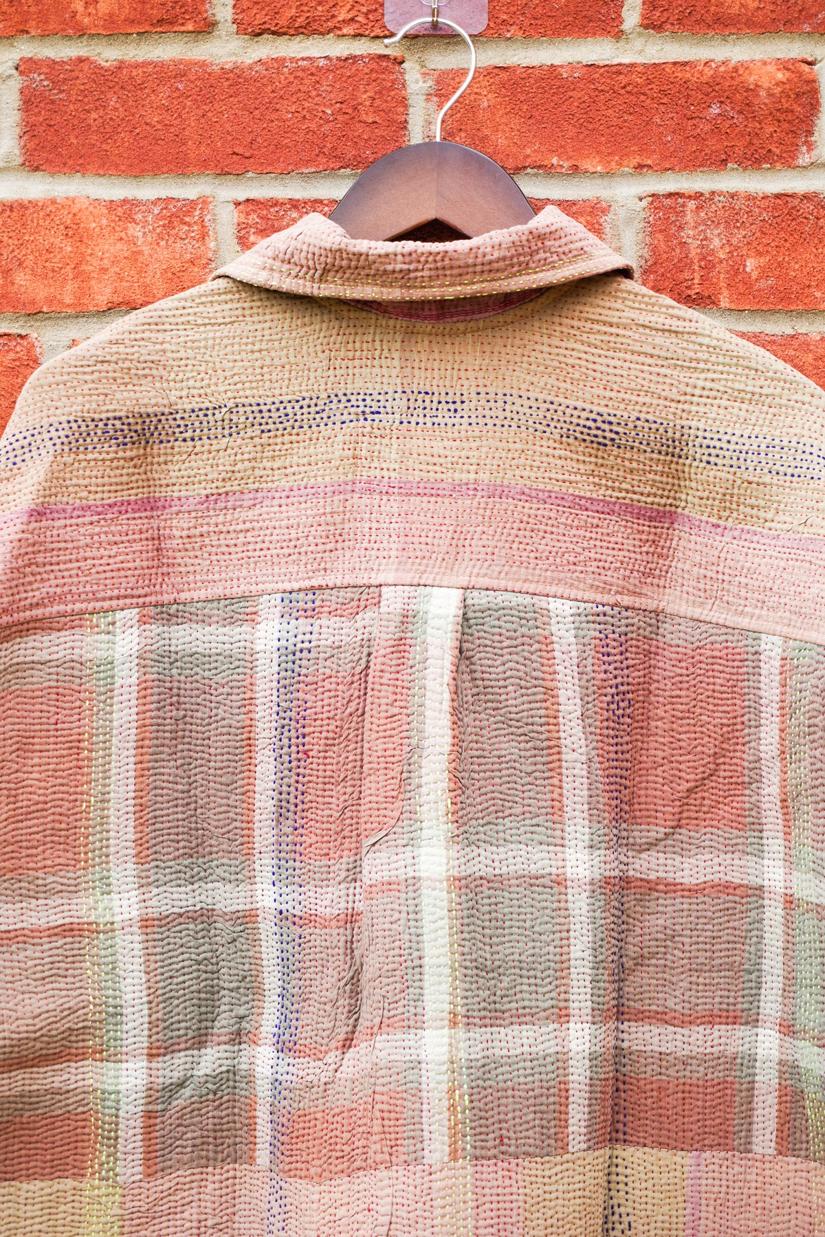 A vintage plaid shirt jacket made from mixed textile scraps, hanging on a wooden hanger against a red brick wall background.