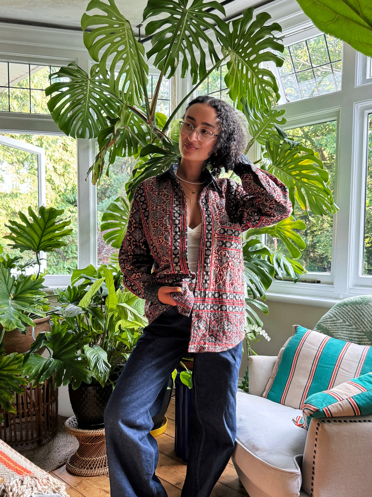 A woman wearing glasses, a patterned button-up Ajrakh Kantha shirt, and dark blue jeans stands in a sunlit room filled with large green plants. 