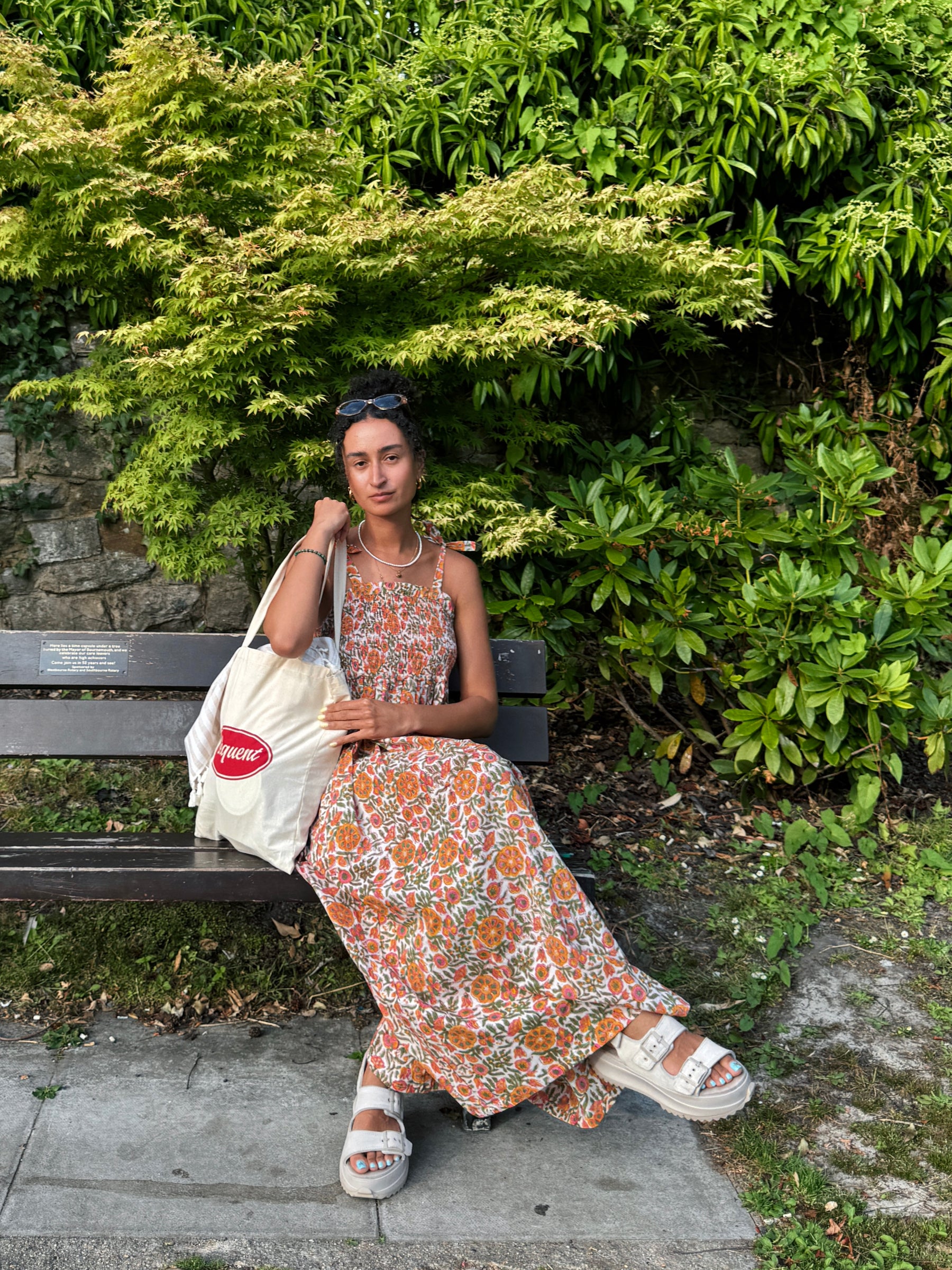 A woman sits on a park bench wearing a sleeveless rust-orange floral hand block painted maxi dress with a white and green pattern, white platform sandals, and carrying a cream tote bag with a red 'Sequent' logo. The background is lush green foliage.