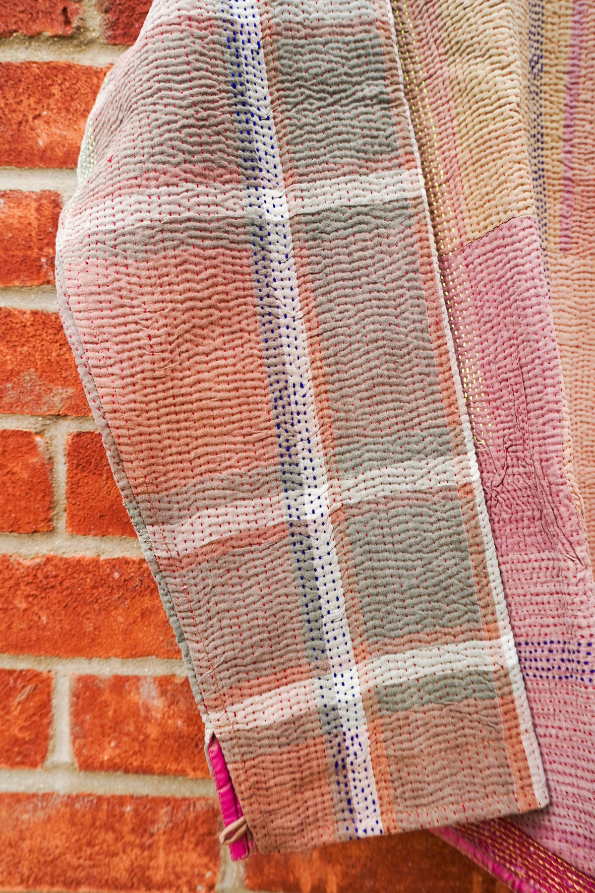 A close-up of a textured, handwoven plaid fabric, likely a scarf or throw blanket, featuring pink, beige, and gray stripes with blue dotted accents, draped against a rustic red brick wall.