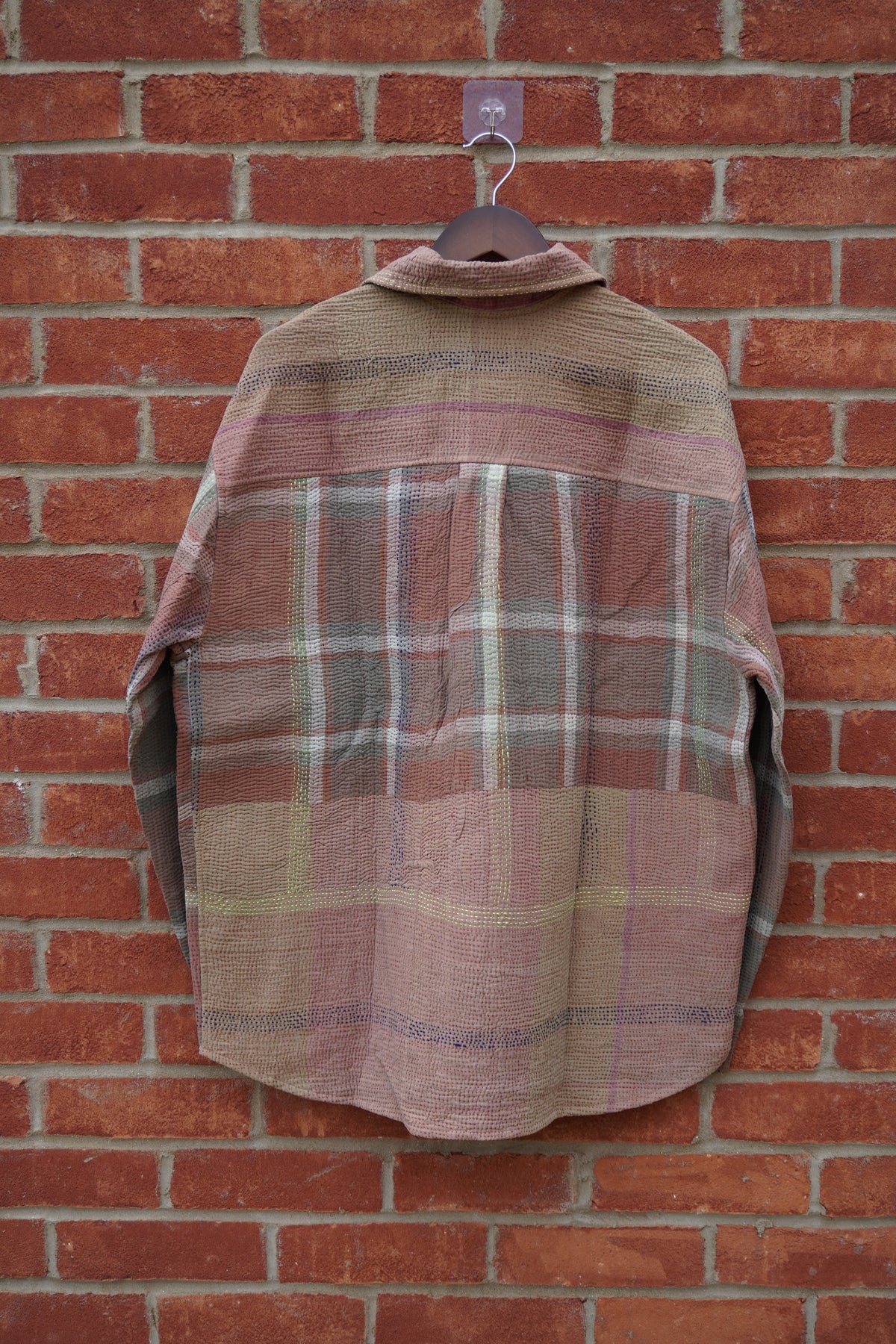 A vintage patchwork plaid flannel shirt in muted earthy tones of pink, brown, and beige, hanging on a wooden hanger against a red brick wall.
