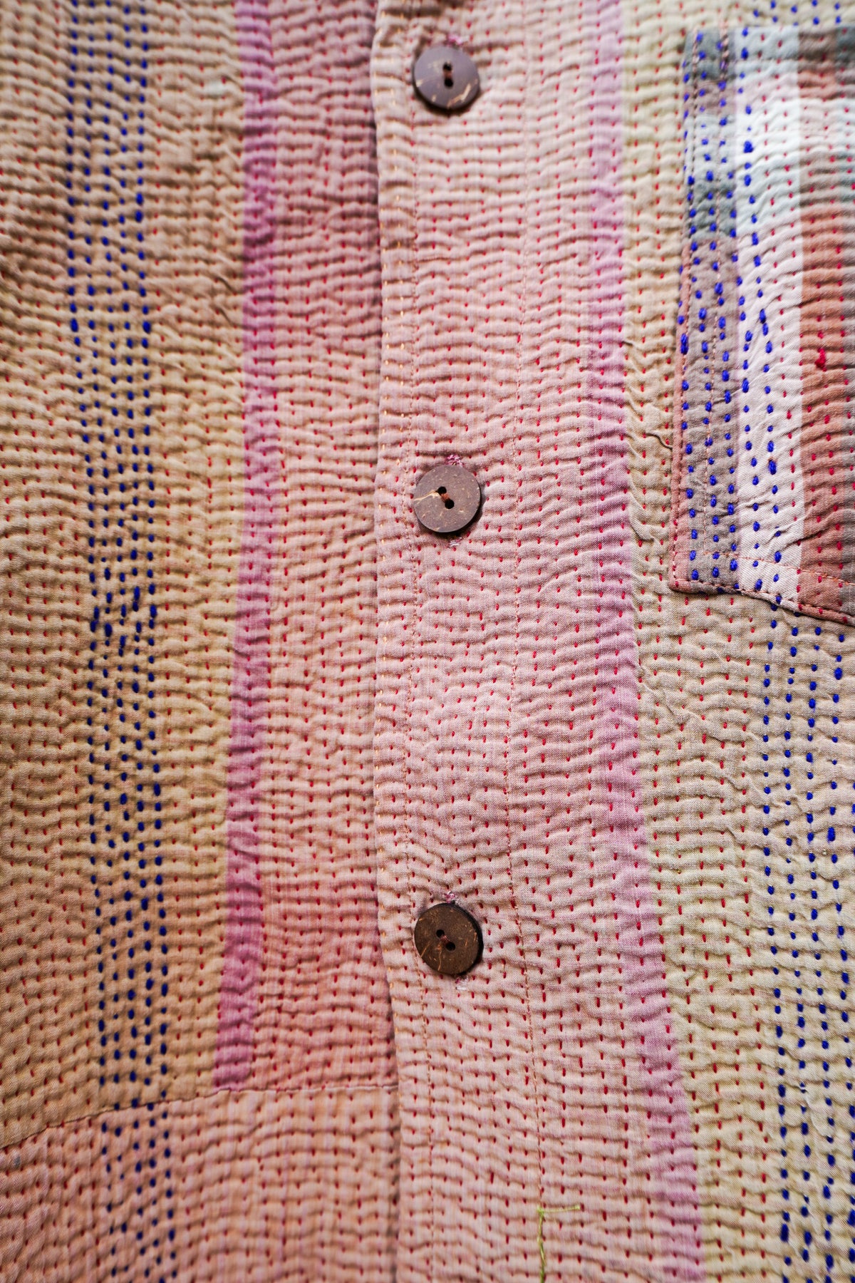 Close-up of a textured woven garment, likely a shirt or jacket, featuring vertical stripes in pink, beige, and blue, with three brown buttons down the front.