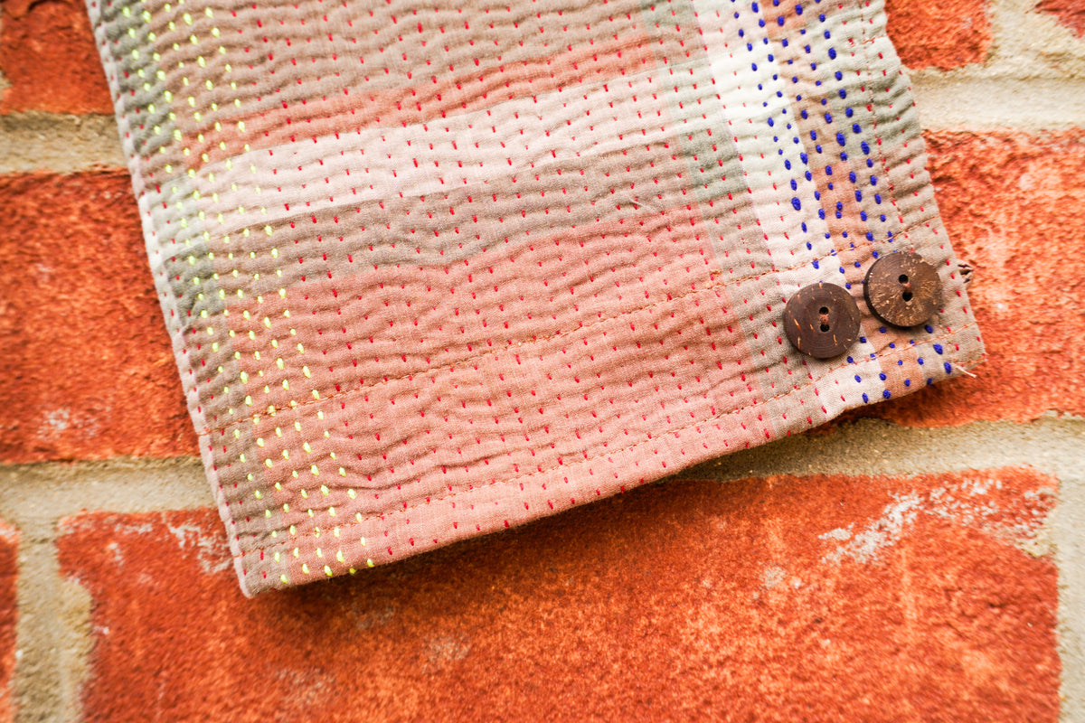 A close-up of a textured, pastel pink and white quilted fabric sleeve with two brown buttons, laid flat on a rustic red-orange brick background.