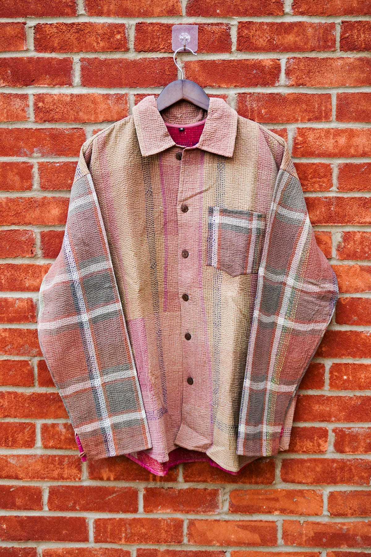 A vintage patchwork plaid button-up shirt in muted earthy tones of pink, beige, and gray, hanging on a wooden hanger against a red brick wall.