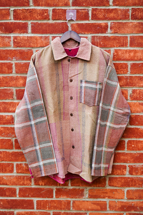 A vintage patchwork plaid button-up shirt in muted earthy tones of pink, beige, and gray, hanging on a wooden hanger against a red brick wall.