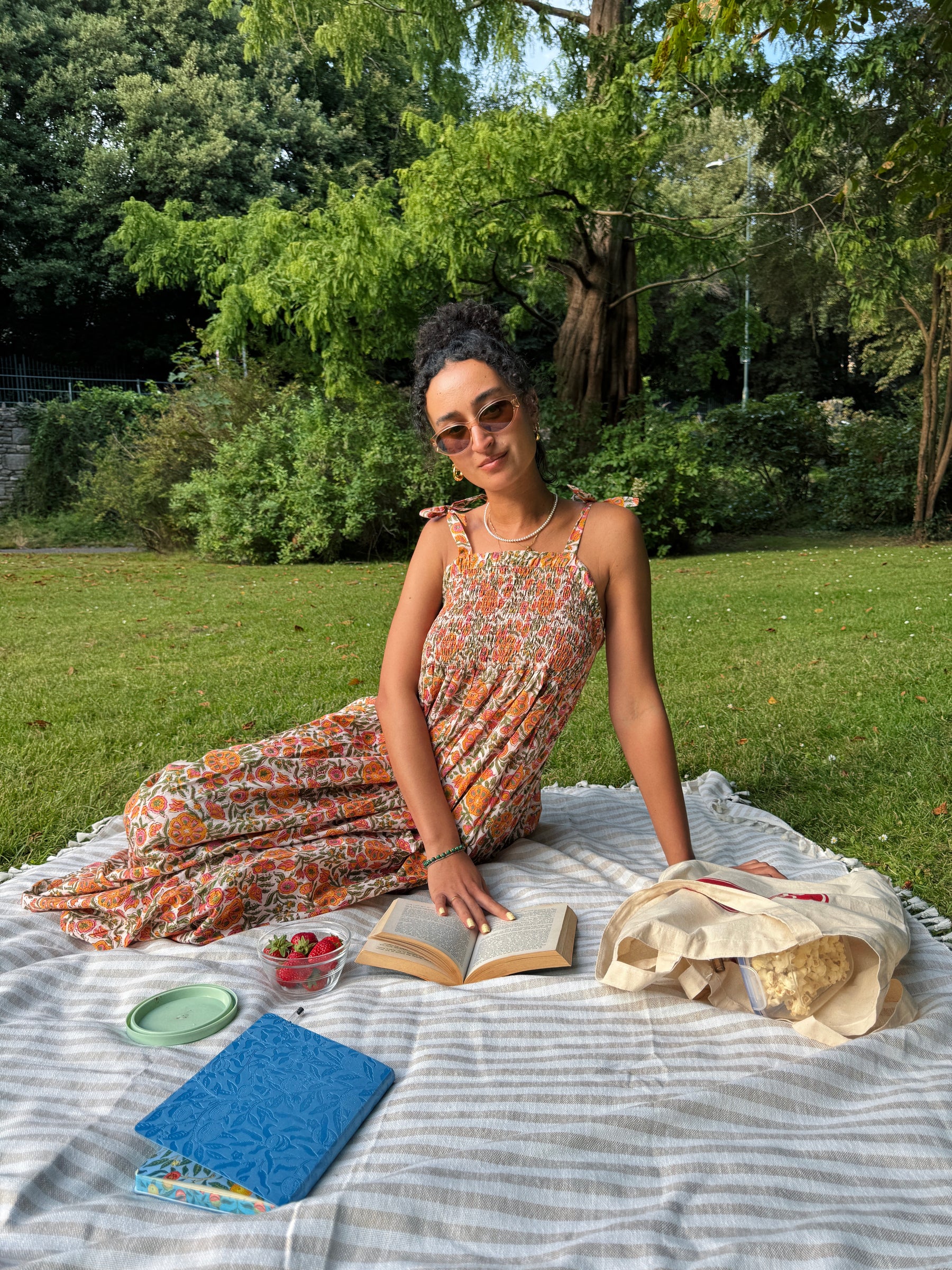 A woman wearing a colorful yellow orange and beige floral hand block painted maxi dress sits on a picnic blanket in a park, surrounded by an open book, a blue notebook, and a canvas tote bag.