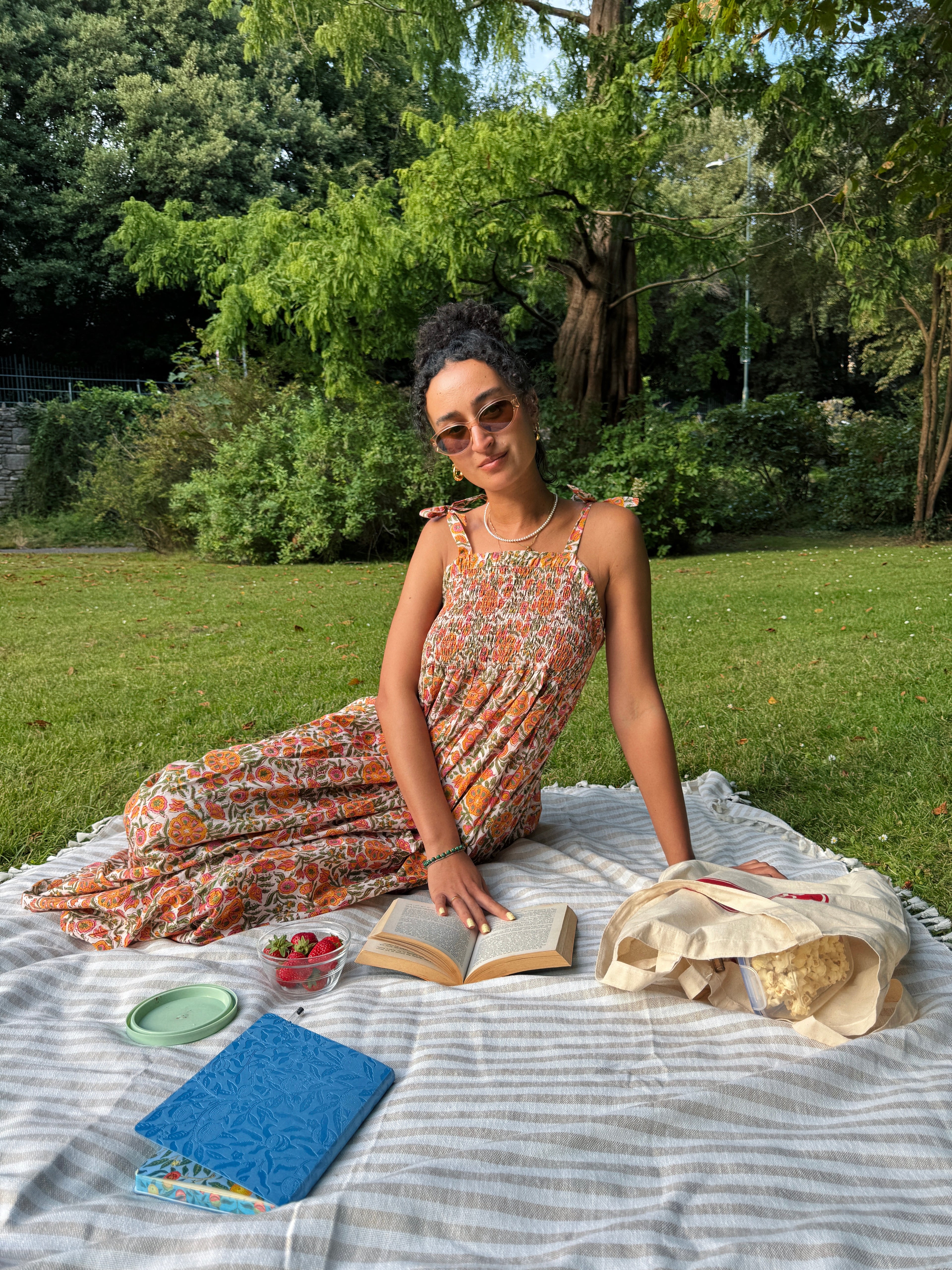 A woman wearing a colorful yellow orange and beige floral hand block painted maxi dress sits on a picnic blanket in a park, surrounded by an open book, a blue notebook, and a canvas tote bag.