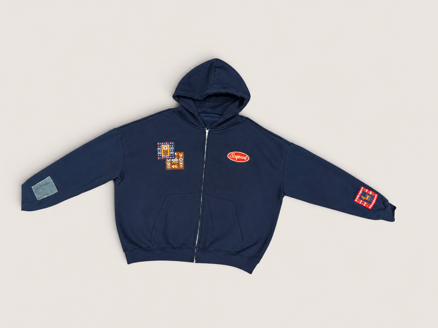 A navy blue zip-up hoodie with multiple colorful embroidered patches, laid flat on a light beige background.