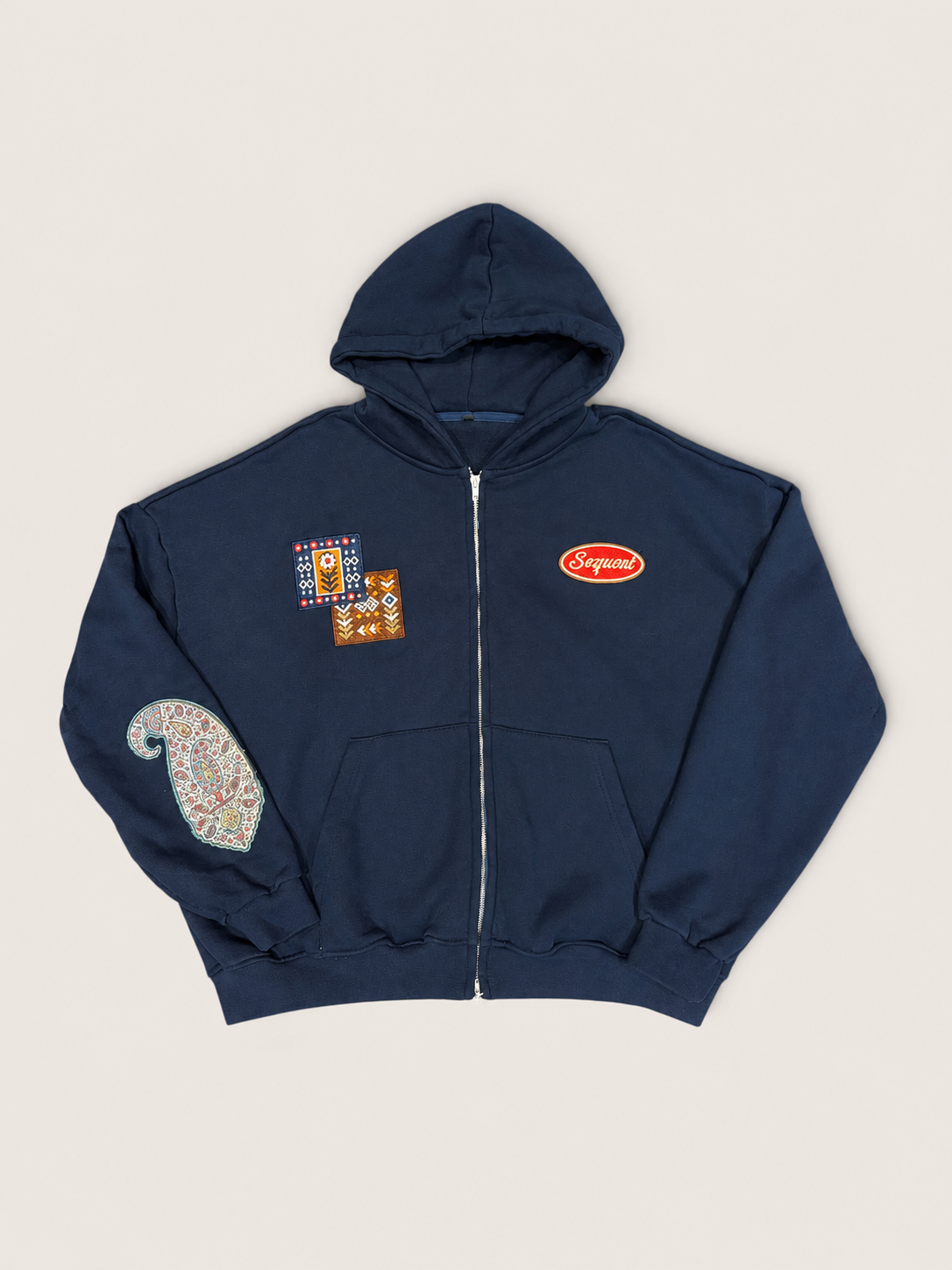 A navy blue hoodie with a double ended front zipper, featuring a paisley patch on the right arm, a small red oval embroidered logo on the left side chest, and a decorative embroidered patch near the left side chest inspired by South Asian architecture, laid flat on a light beige background.