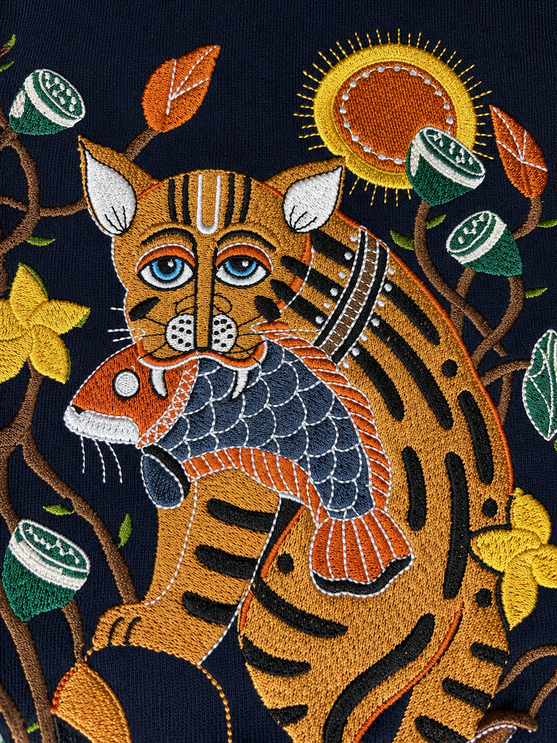 Close-up of a dark navy embroidered textile featuring a tiger holding a fish, with a yellow sun, orange leaves, and green and yellow flowers.
