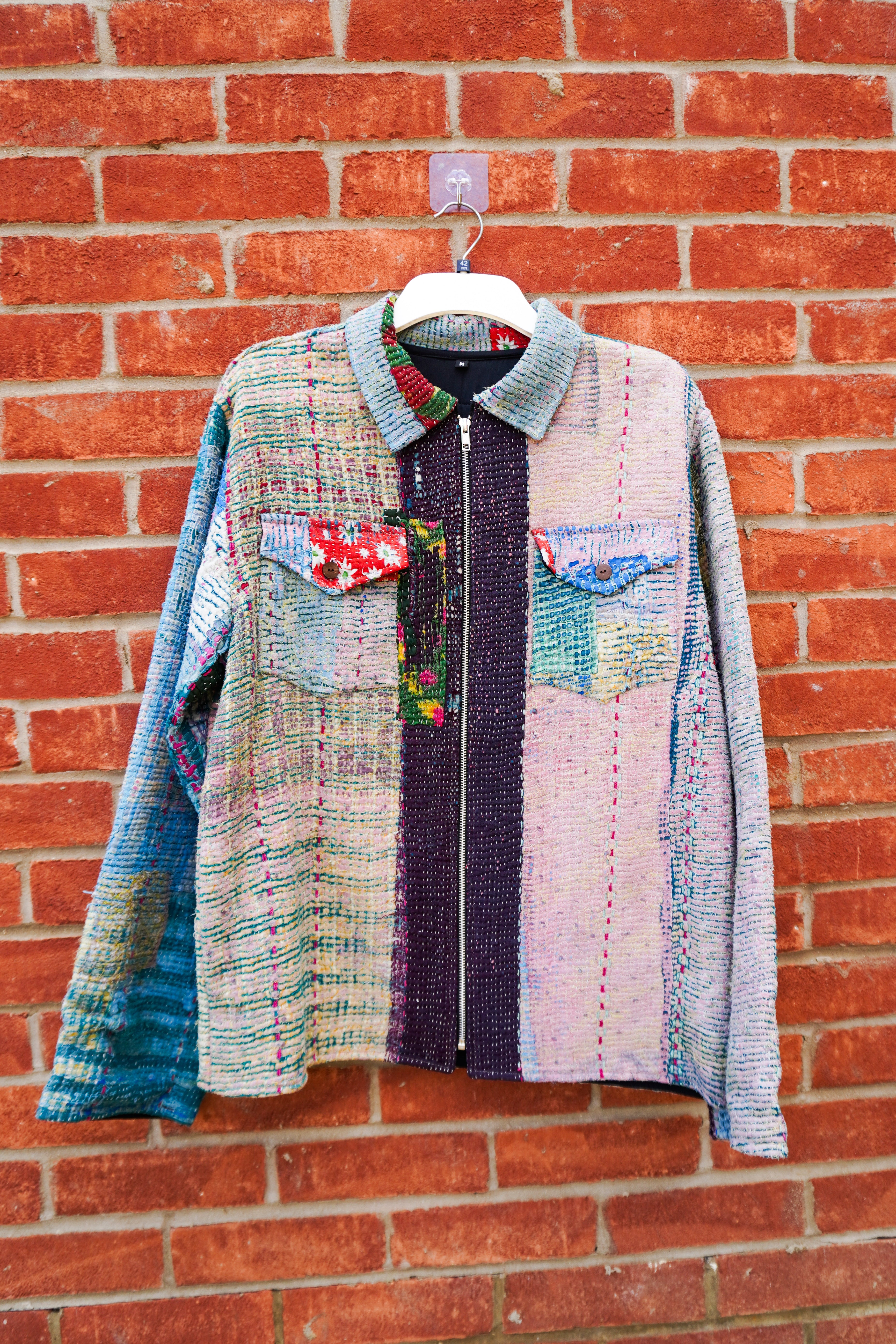 A long-sleeve zip-front jacket made from patchwork woven kantha textile panels in blue, pink, purple, green, and beige, hanging on a white hanger against a red brick wall.