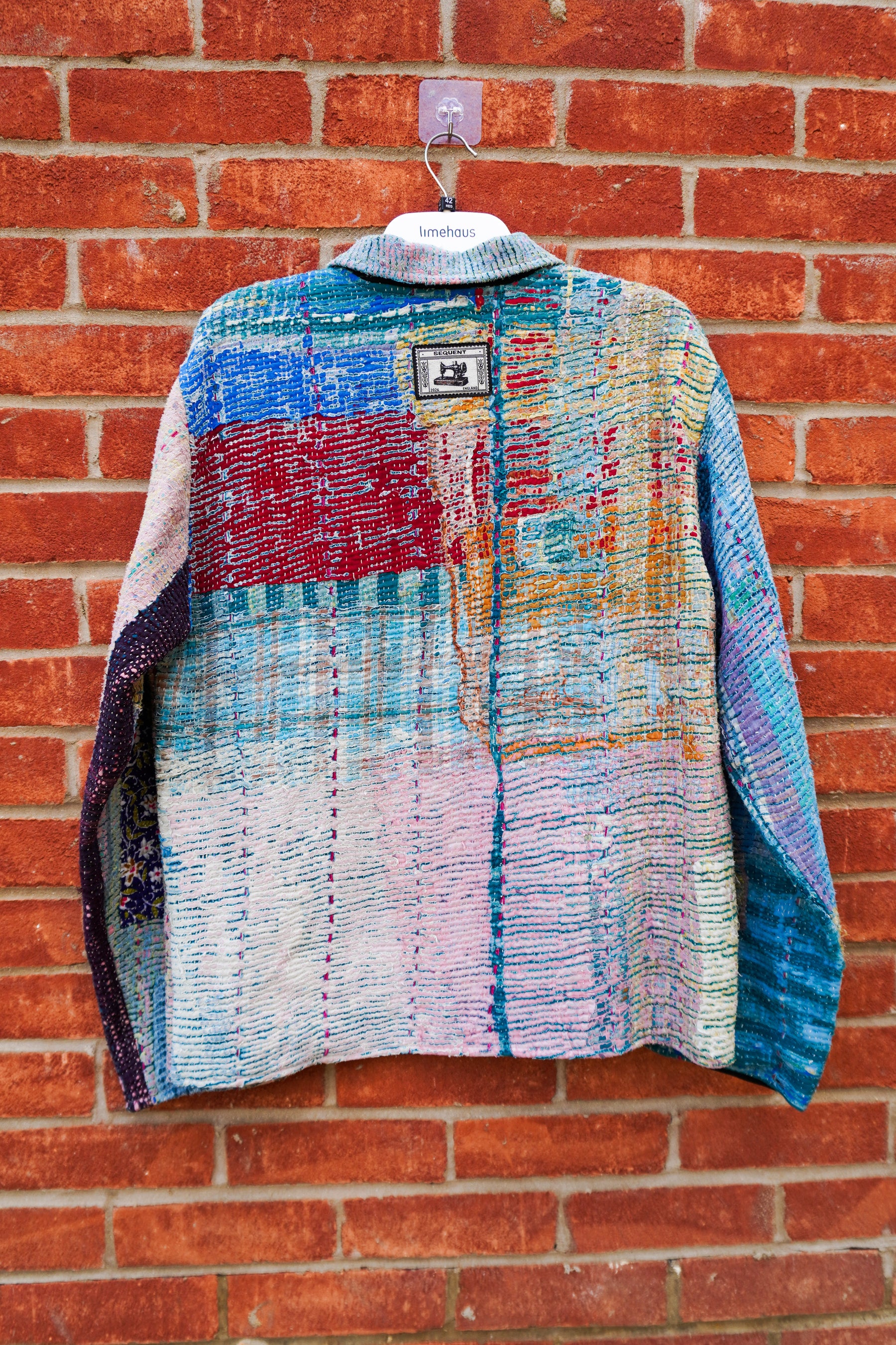 A multicolor patchwork knit kantha jacket from behind with a textured, handwoven appearance, hanging on a white hanger against a red brick wall.
