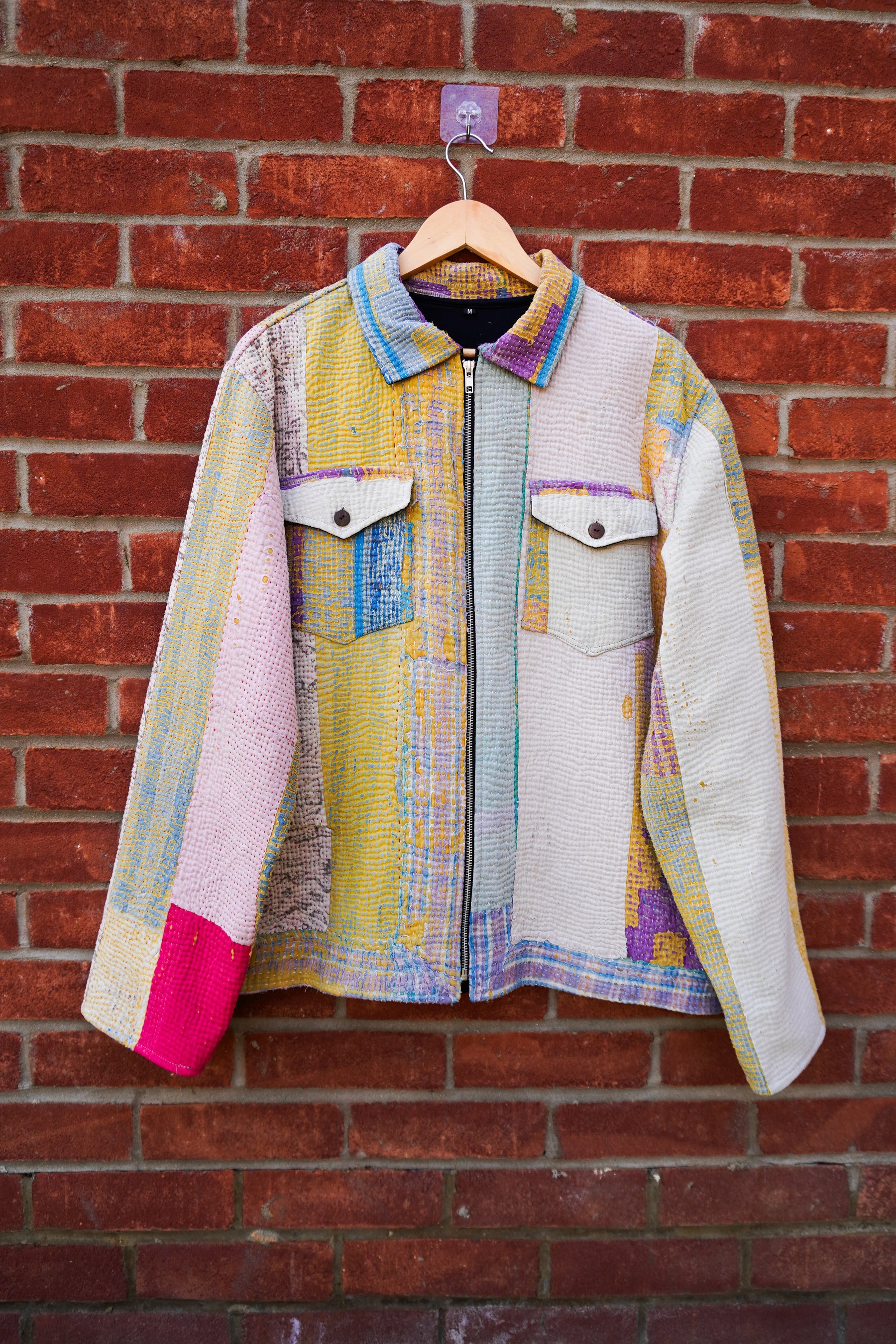 A multicolor patchwork quilted kantha jacket hanging on a wooden hanger against a red brick wall. The jacket features a mix of pastel and bright colors including yellow, pink, blue, purple, and white in a quilted pattern, with a black zipper and two buttoned chest pockets.