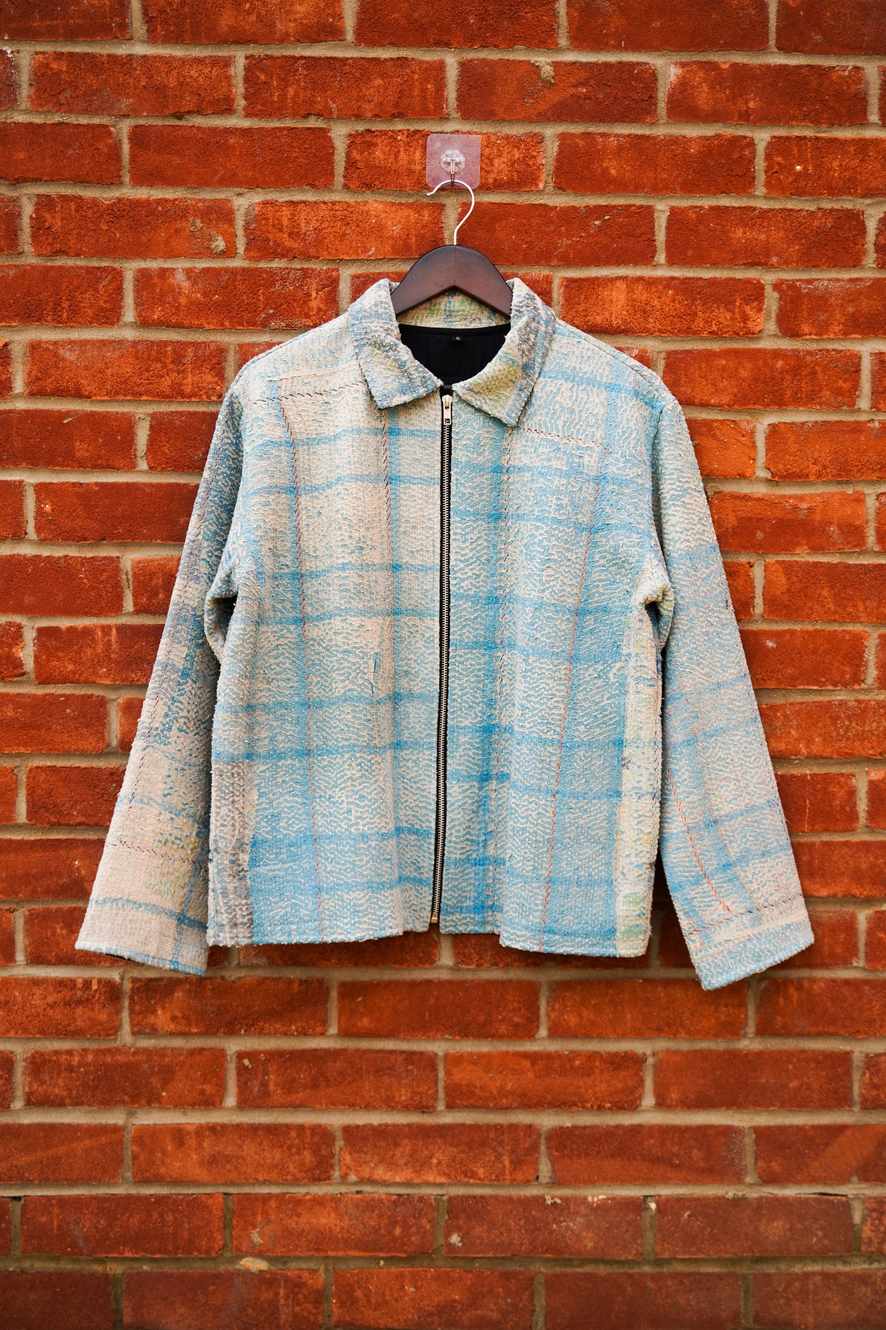 A light blue and beige plaid zip-up kantha jacket hanging on a dark wooden hanger against a red brick wall.