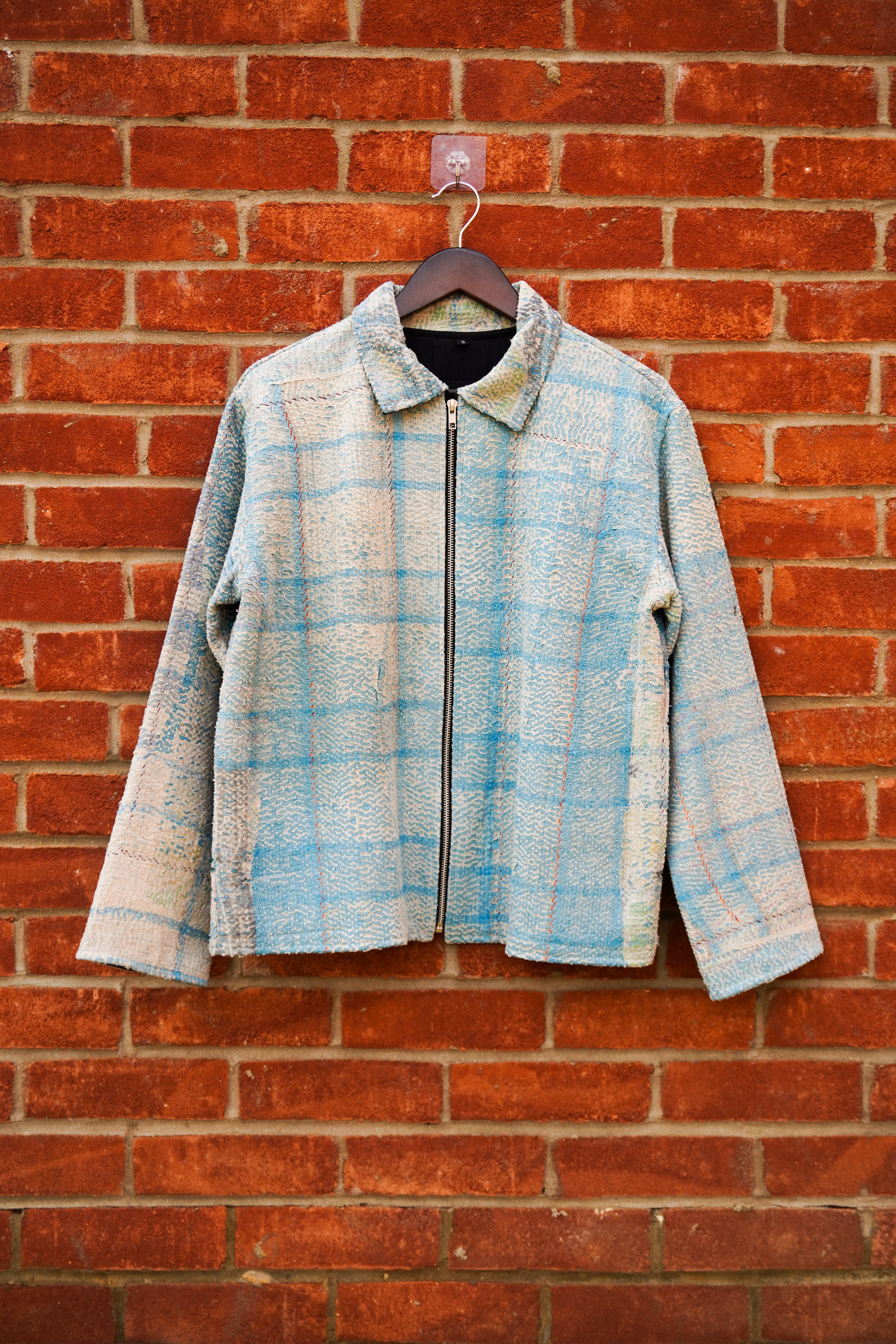 A light blue and beige plaid zip-up kantha jacket hanging on a dark wooden hanger against a red brick wall.