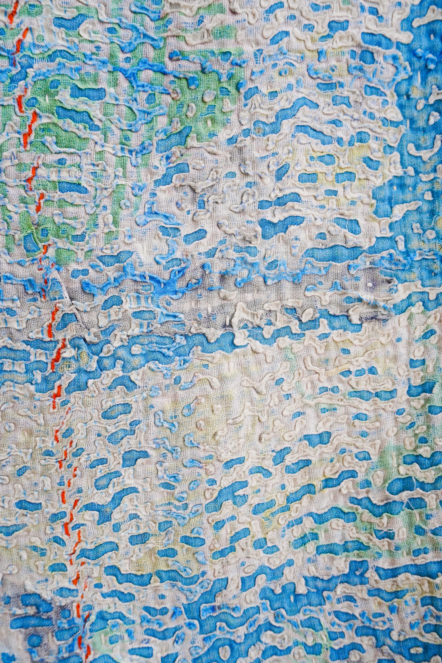 A close-up of a textured kantha fabric featuring organic wavy shapes in shades of blue, green, and red on a cream and beige background.