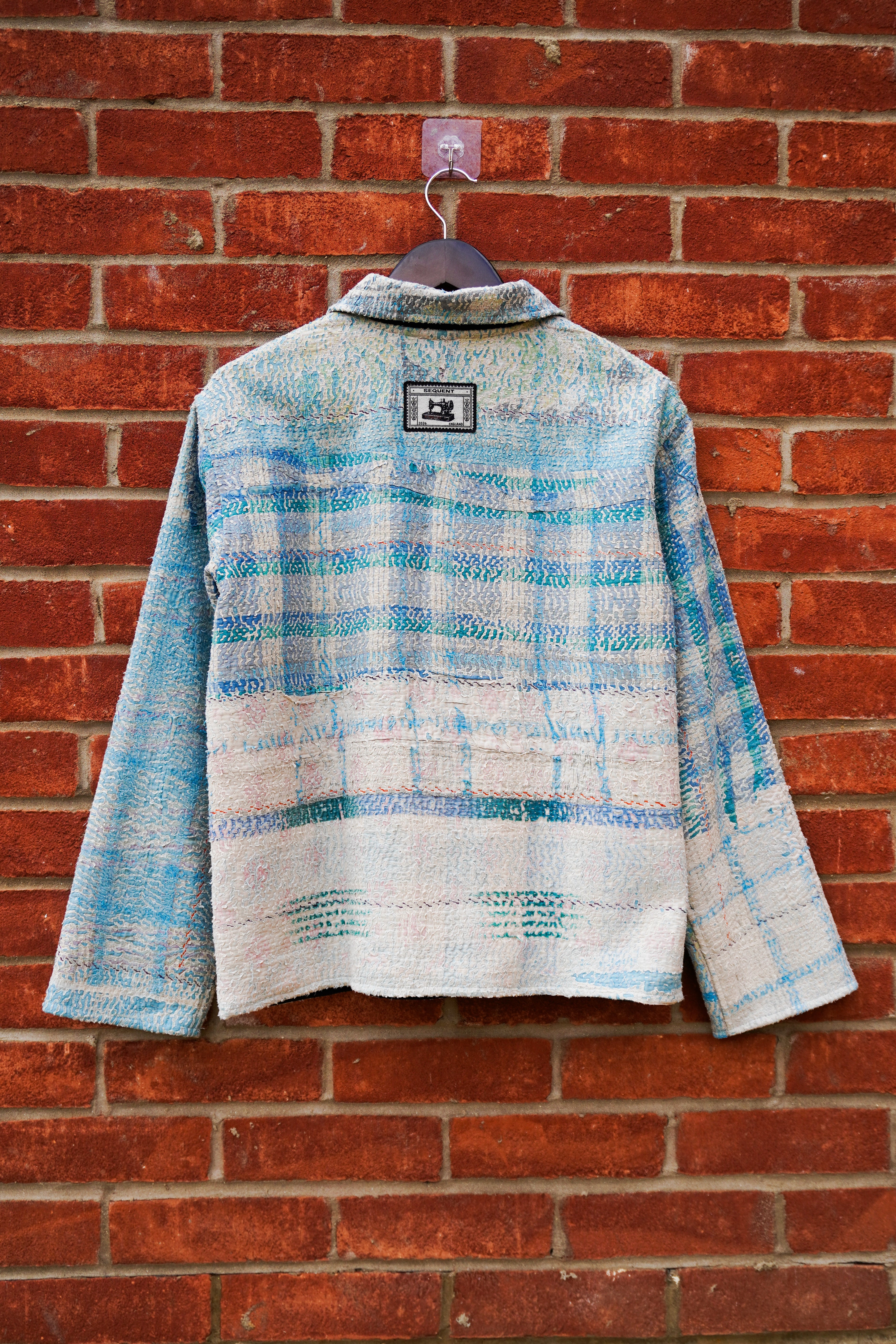 A textured, long-sleeve cropped kantha jacket with a blue and beige plaid pattern, hanging on a dark hanger against a red brick wall.