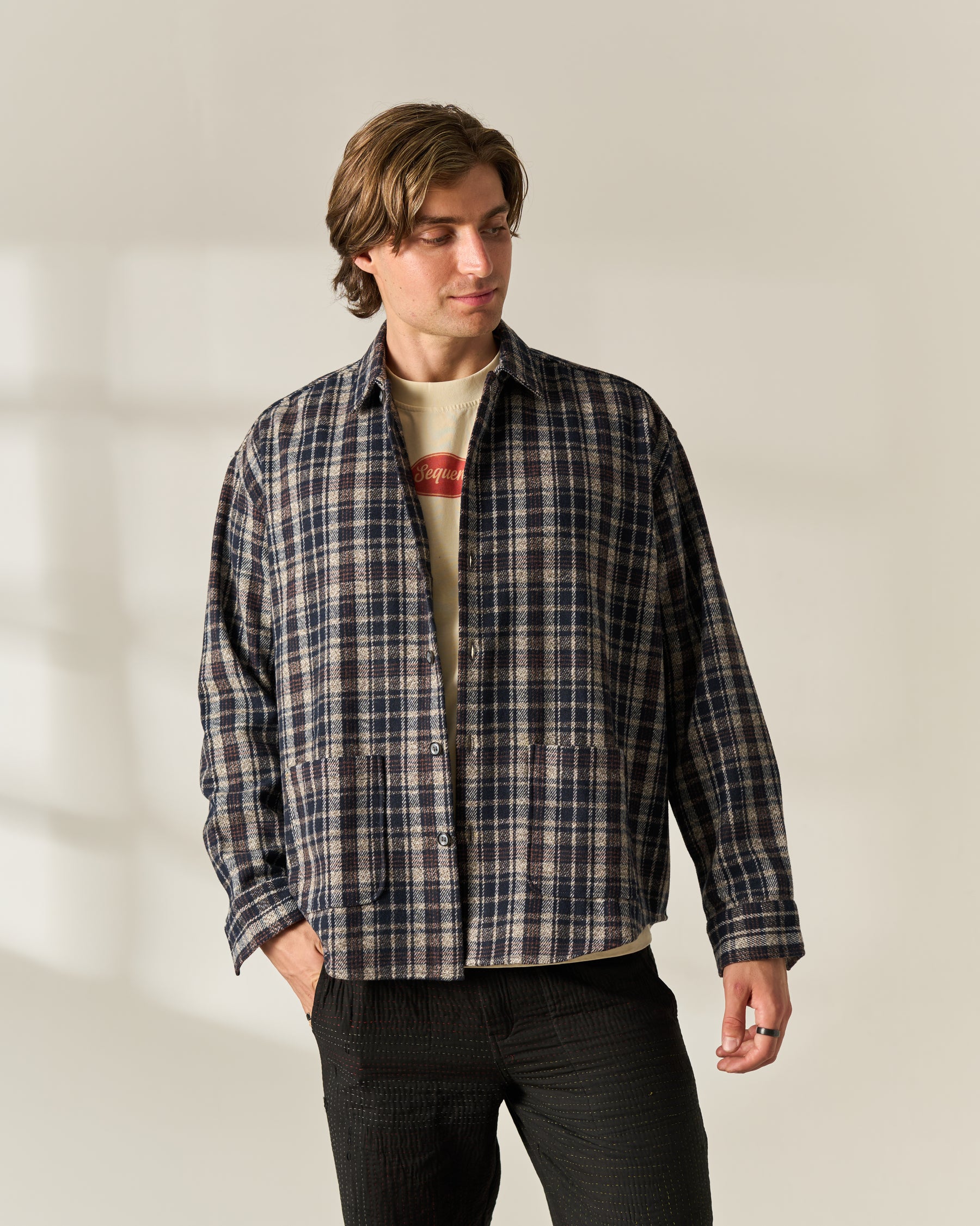 A man wearing a dark navy and beige plaid wool blend overshirt over a cream graphic t-shirt and dark charcoal pants, standing against a warm off-white studio background with soft window shadow details.