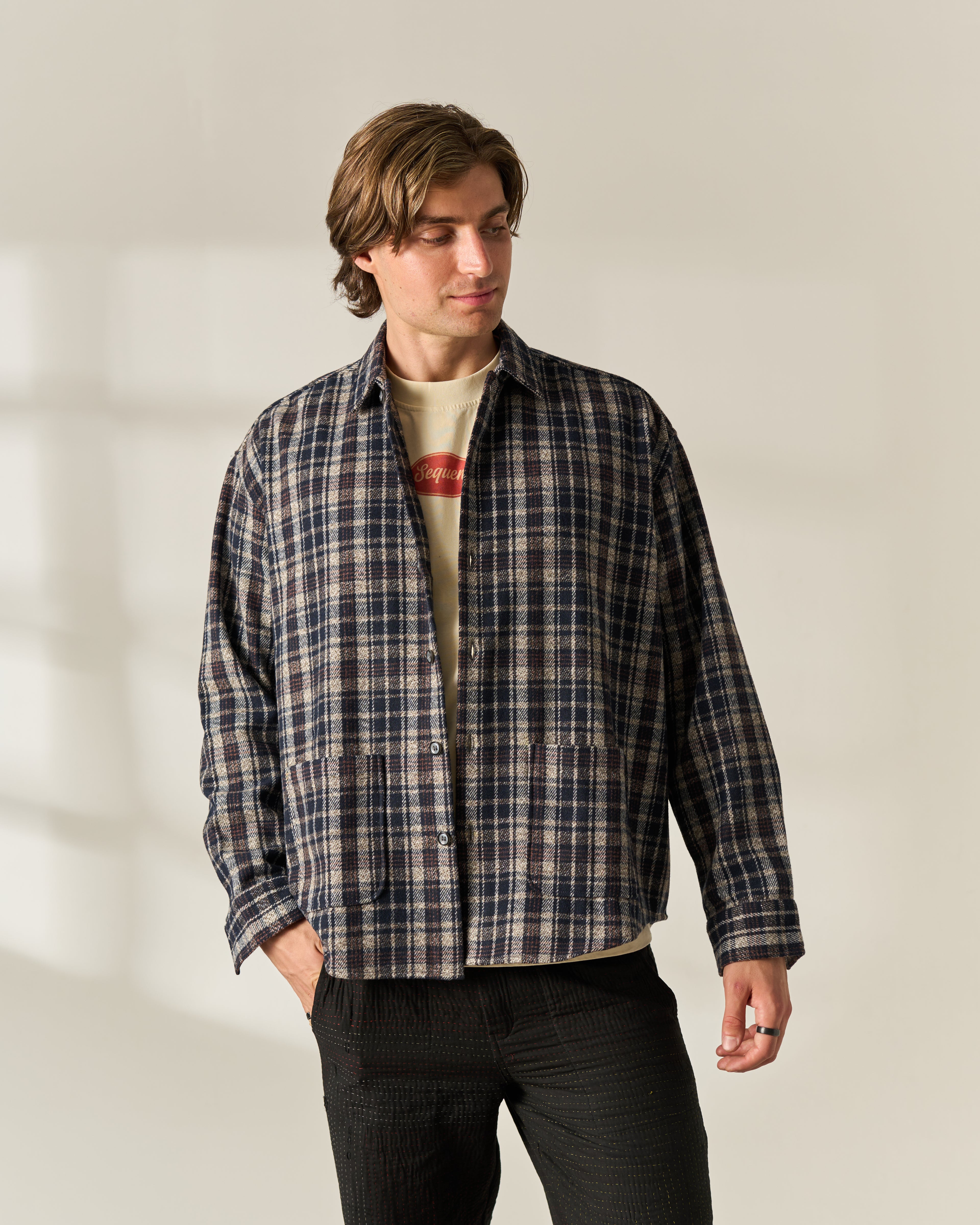 A man wearing a dark navy and beige plaid wool blend overshirt over a cream graphic t-shirt and dark charcoal pants, standing against a warm off-white studio background with soft window shadow details.
