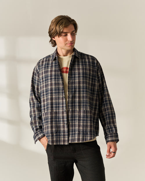 A man wearing a dark navy and beige plaid wool blend overshirt over a cream graphic t-shirt and dark charcoal pants, standing against a warm off-white studio background with soft window shadow details.