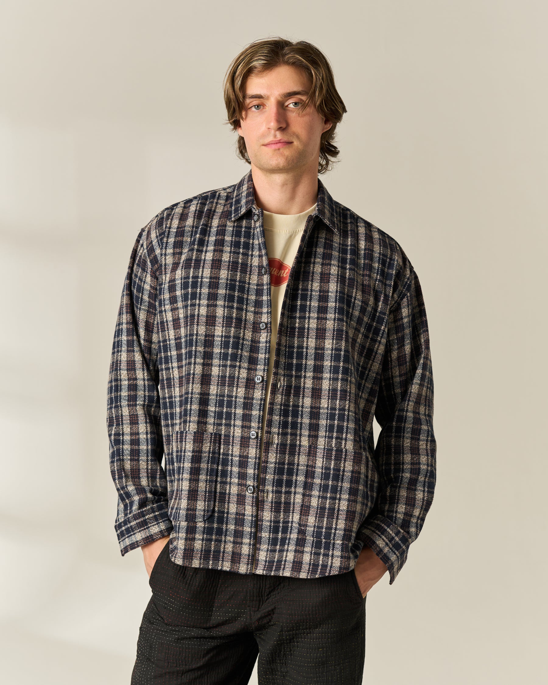 A man wearing a navy, beige, and brown plaid button-up wool blend Over-shirt over a cream graphic t-shirt and dark pants, standing against a plain beige studio background.