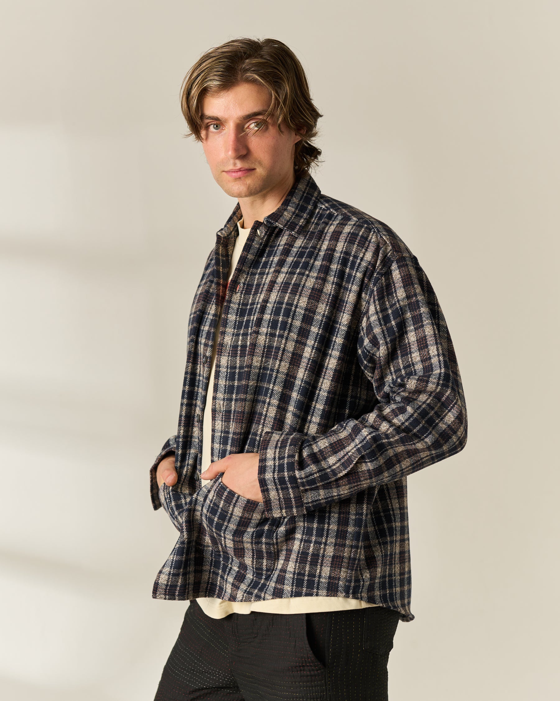 A man wearing a long-sleeve navy and beige plaid button-down wool blend overshirt over a cream tshirt, paired with dark pants, standing against a plain light beige background.