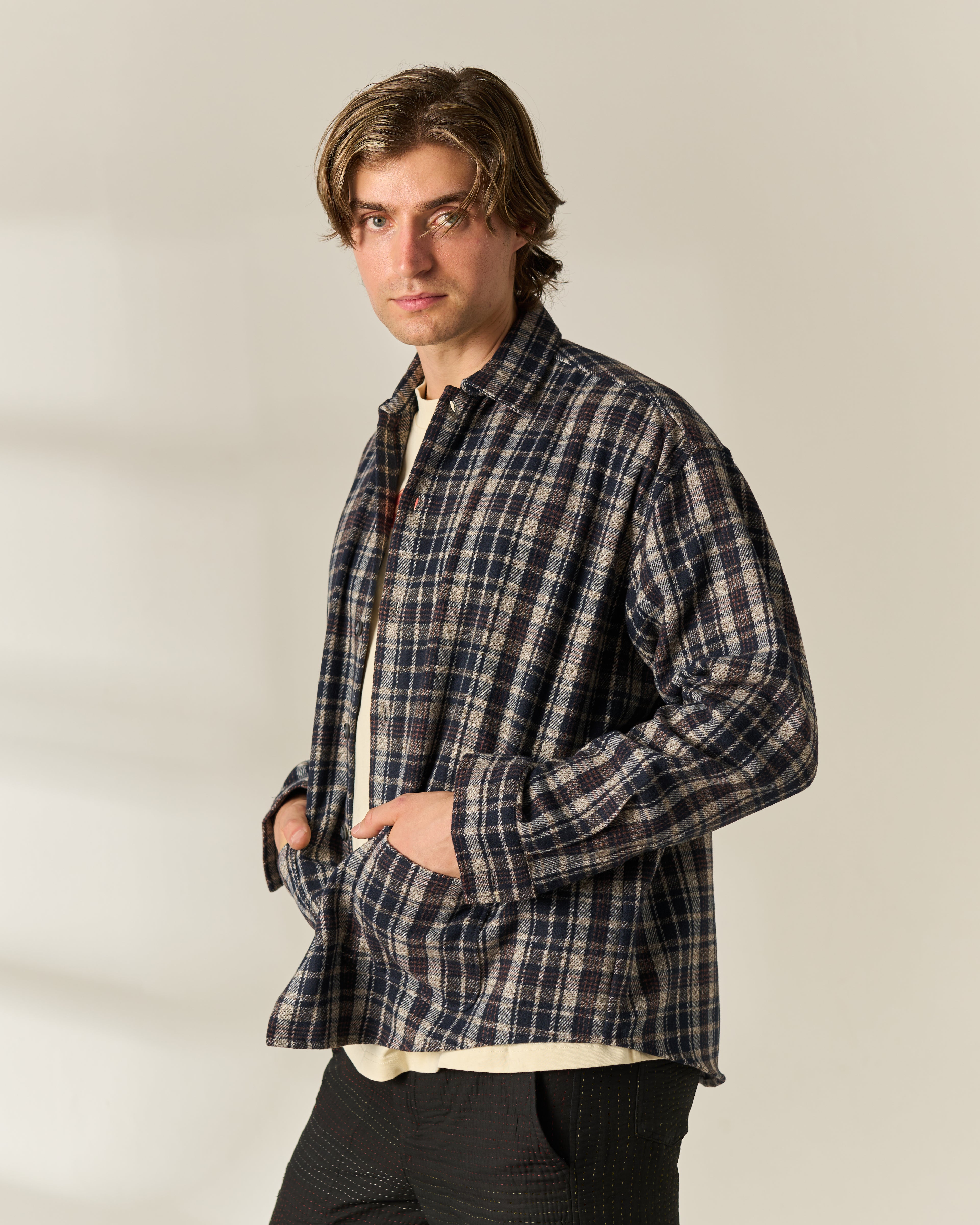 A man wearing a long-sleeve navy and beige plaid button-down wool blend overshirt over a cream tshirt, paired with dark pants, standing against a plain light beige background.