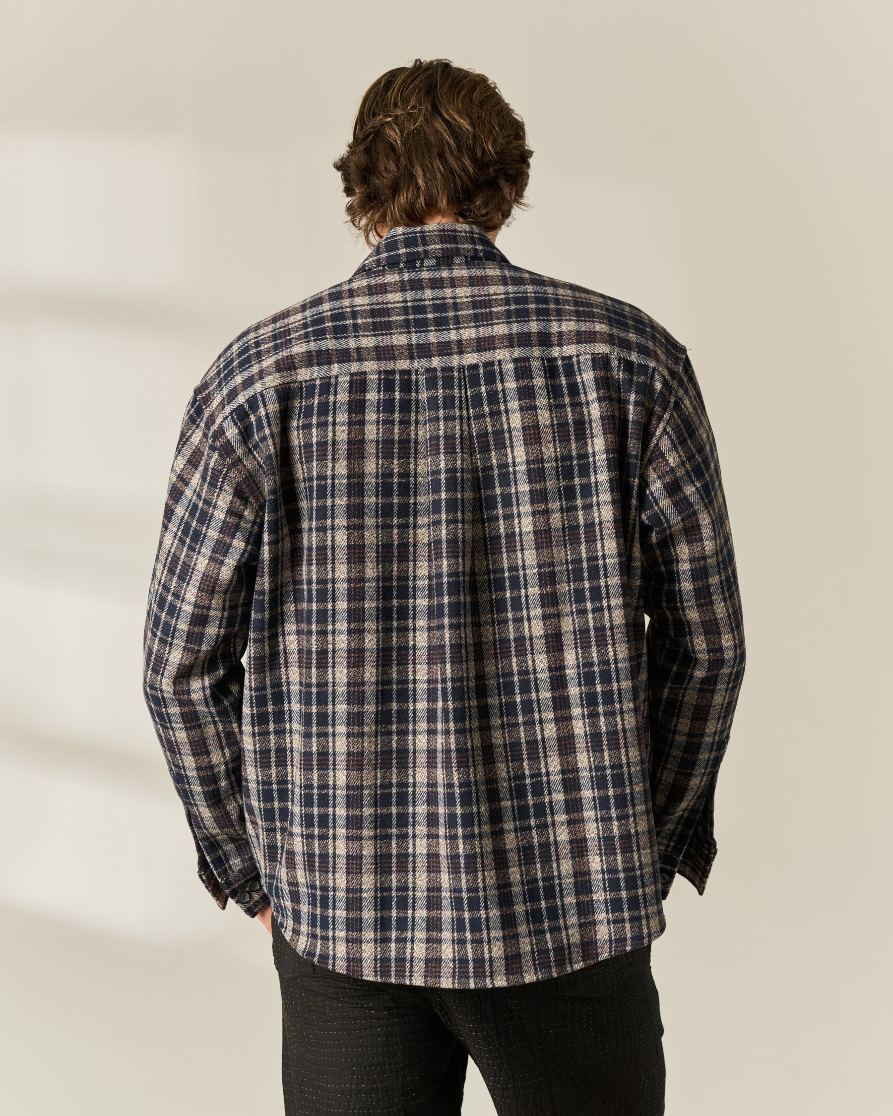 A man viewed from behind wearing a long-sleeve plaid button-up wool blend Overshirt in navy, beige, and white, paired with dark pants, against a light beige background.
