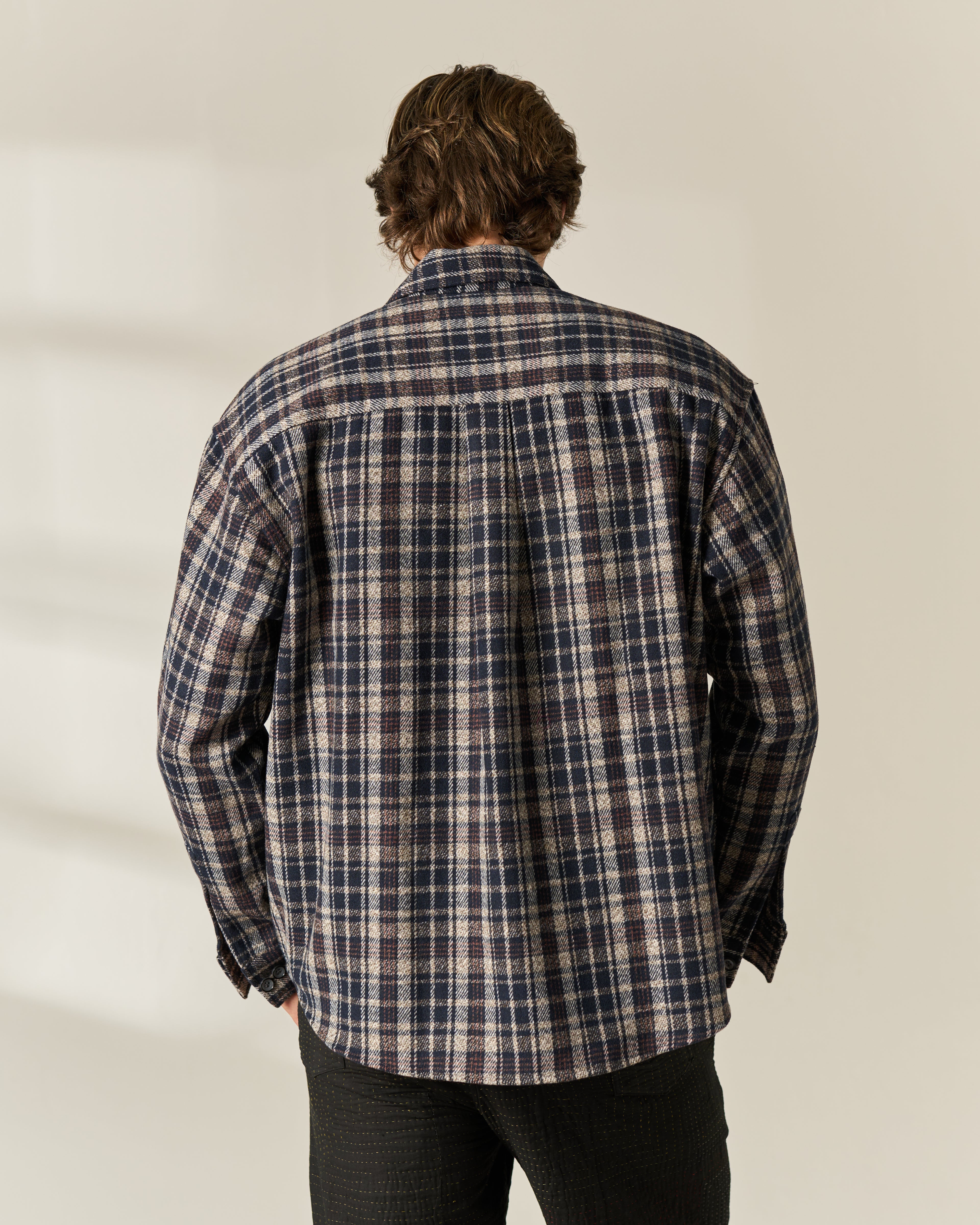 A man viewed from behind wearing a long-sleeve plaid button-up wool blend Overshirt in navy, beige, and white, paired with dark pants, against a light beige background.
