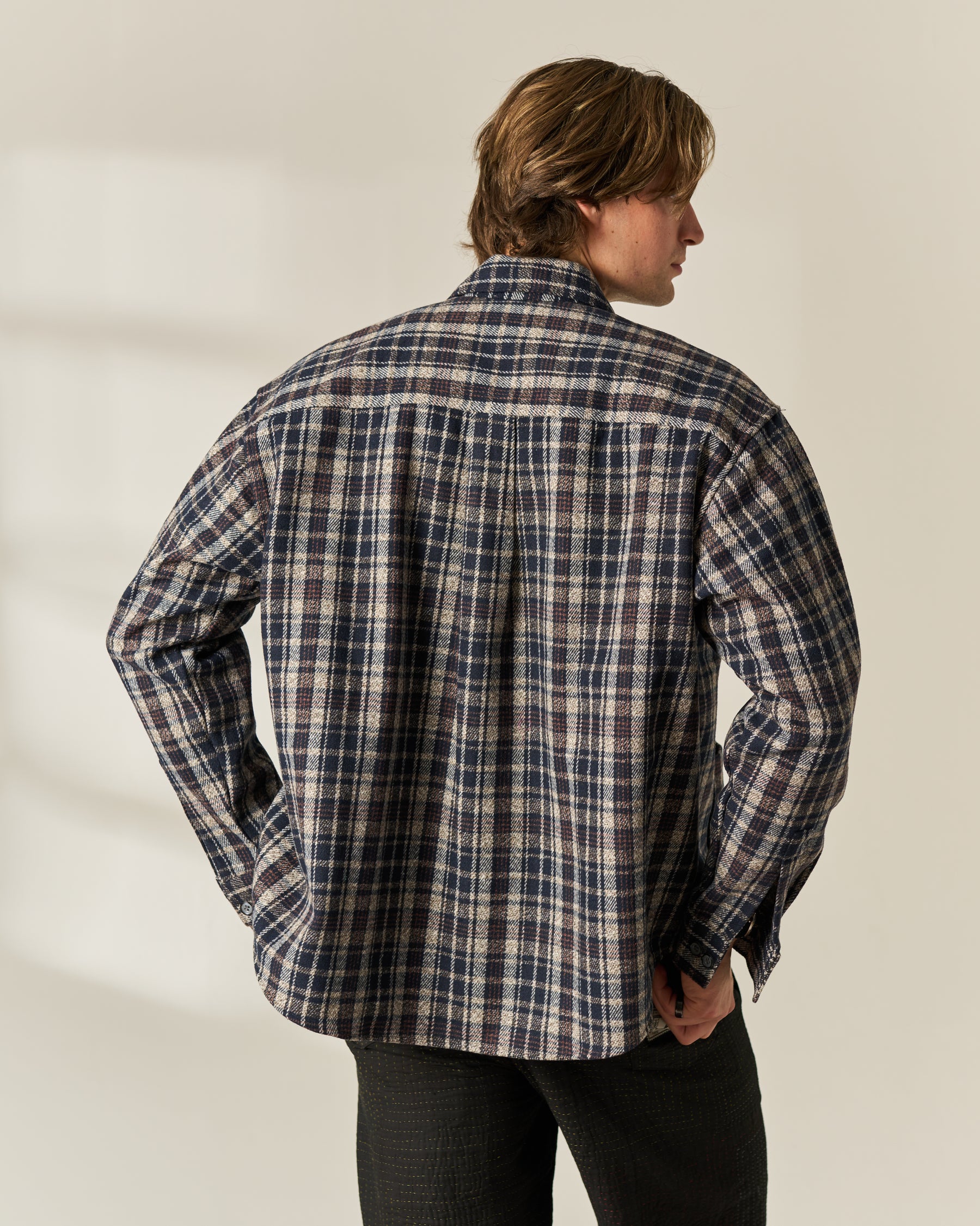 A man shown from the back wearing a long-sleeve plaid button-up wool blend overshirt in navy, beige, and white, paired with dark pants, against a light beige studio background.