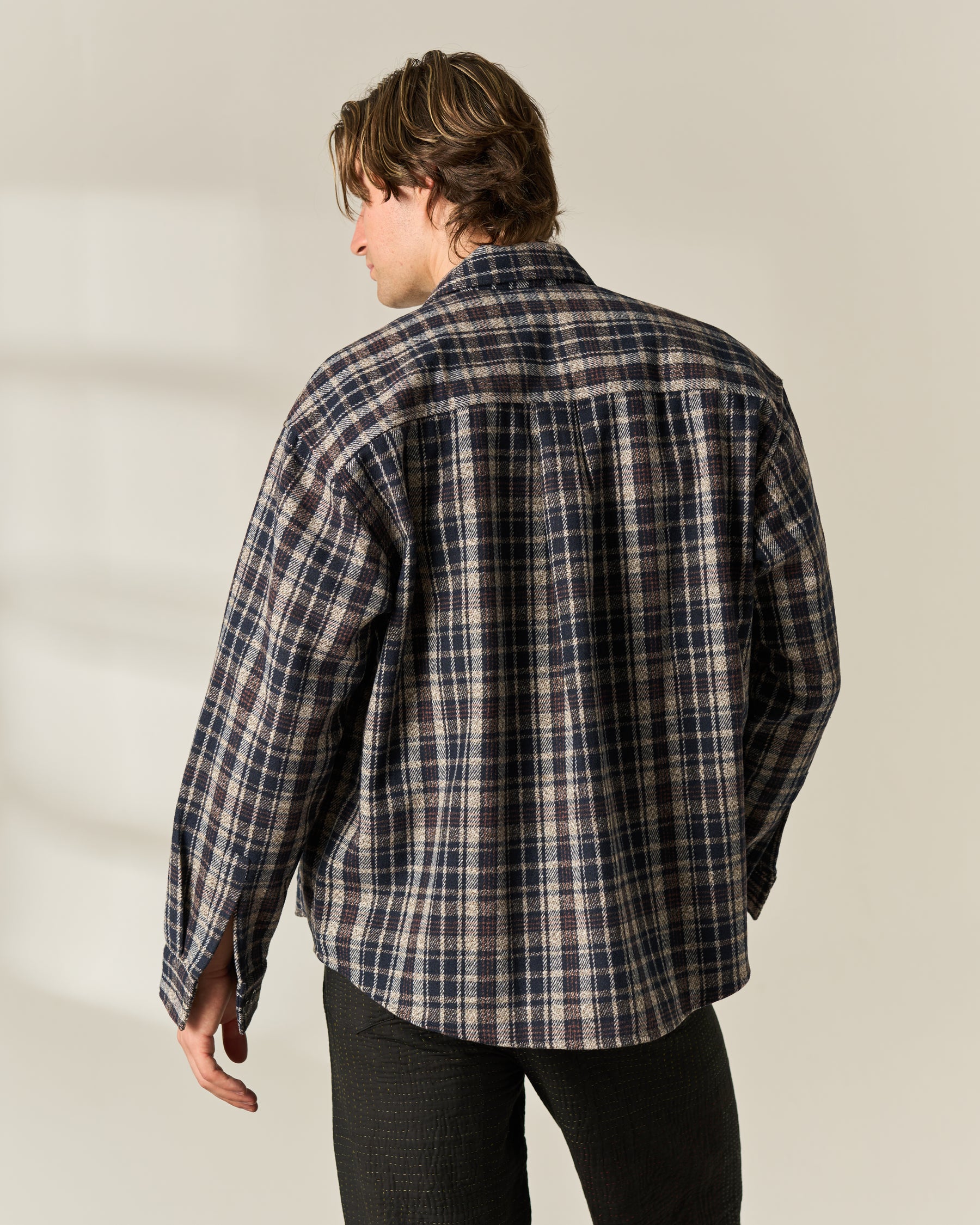 A man shown from behind wearing a long-sleeve plaid button-up wool blend Over-shirt in navy, beige, and gray, paired with dark pants, against a plain light beige background.