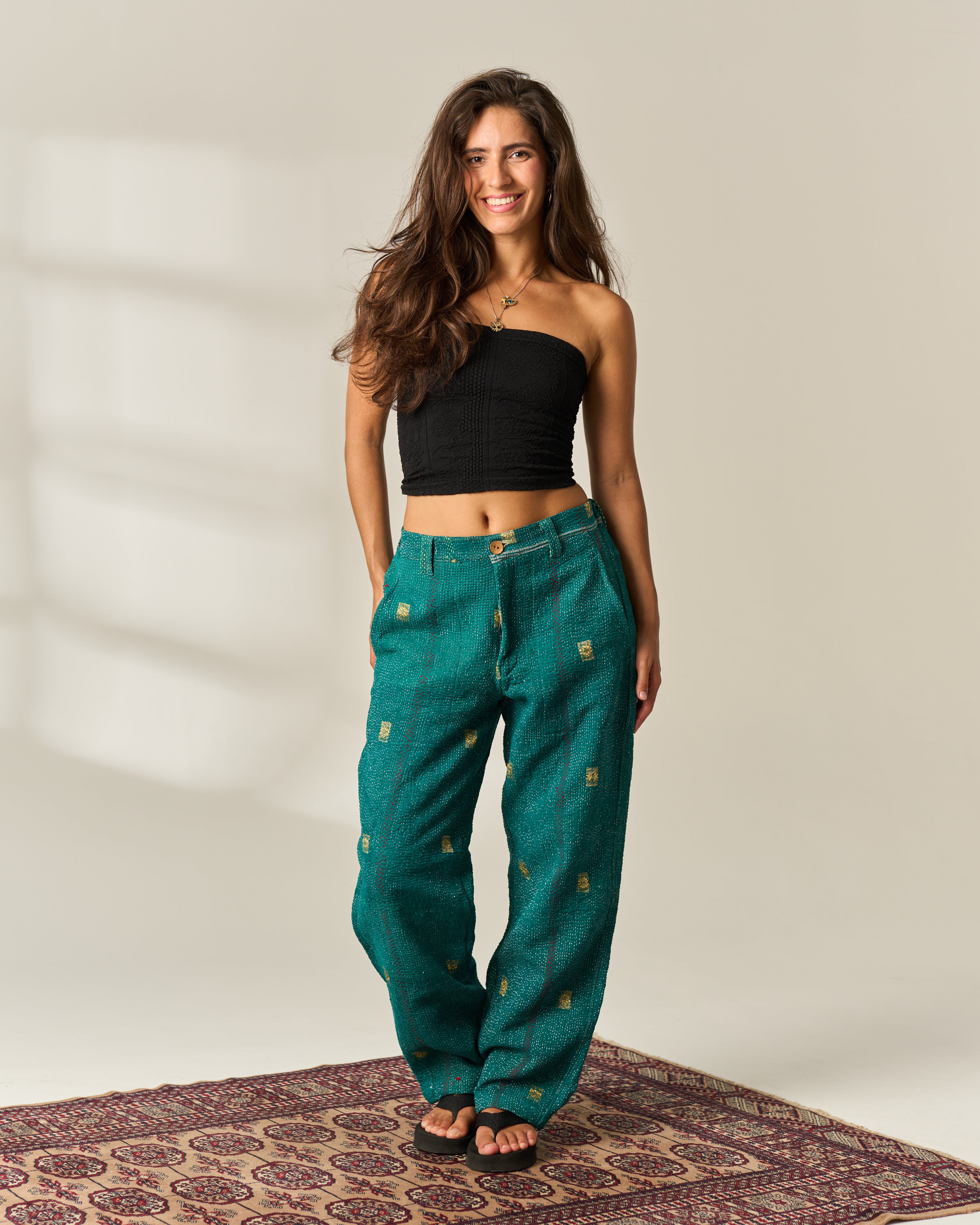 A woman stands on a patterned rug wearing a black strapless crop top and wide-leg teal-green vintage Kantha pants with small gold embroidered motifs, hand-stitched detailing, photographed in a softly lit studio setting.