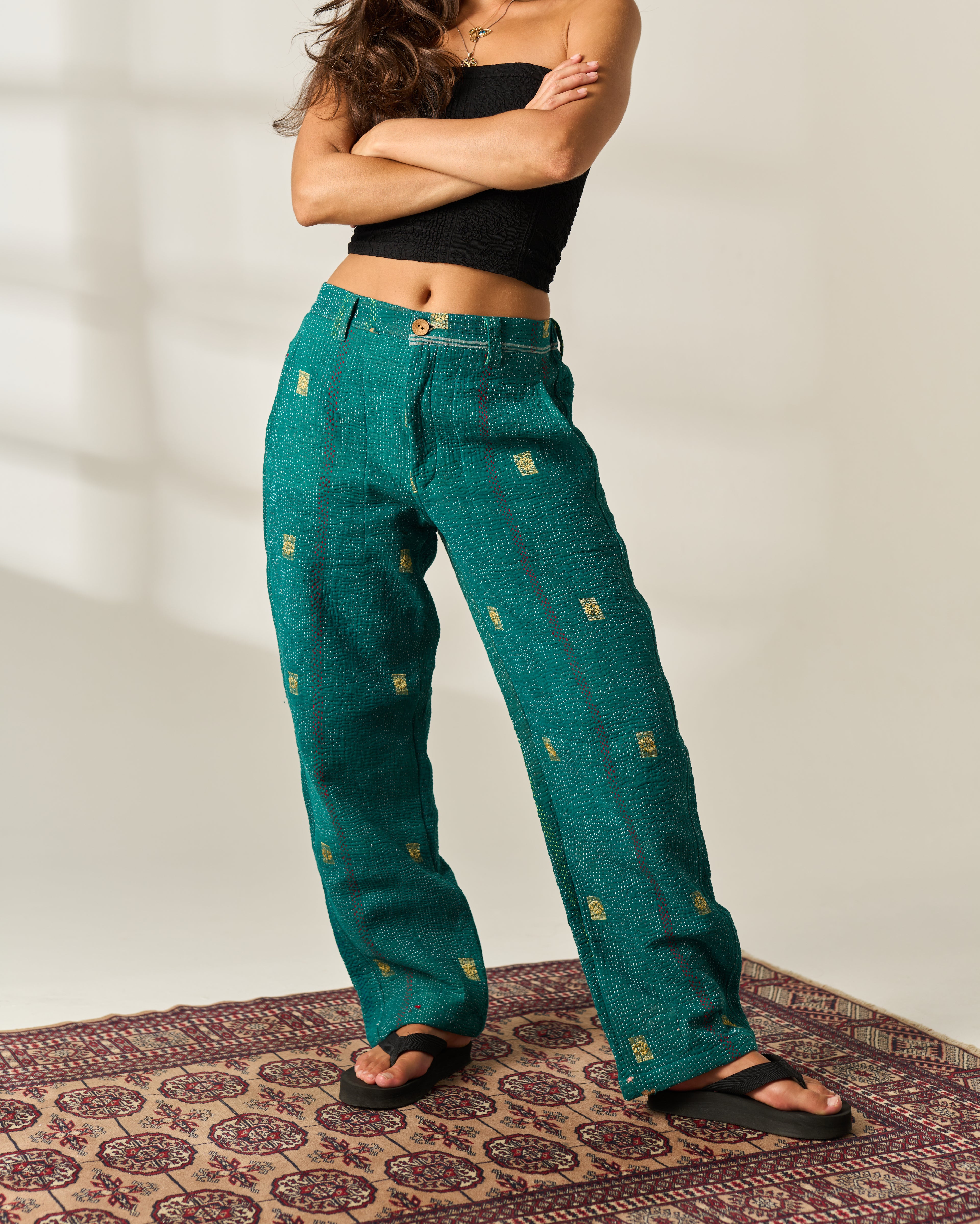 A model wearing teal green wide-leg vintage Kantha trousers with small gold hand embroidered details, styled with a black crop top and black sandals on a patterned rug.