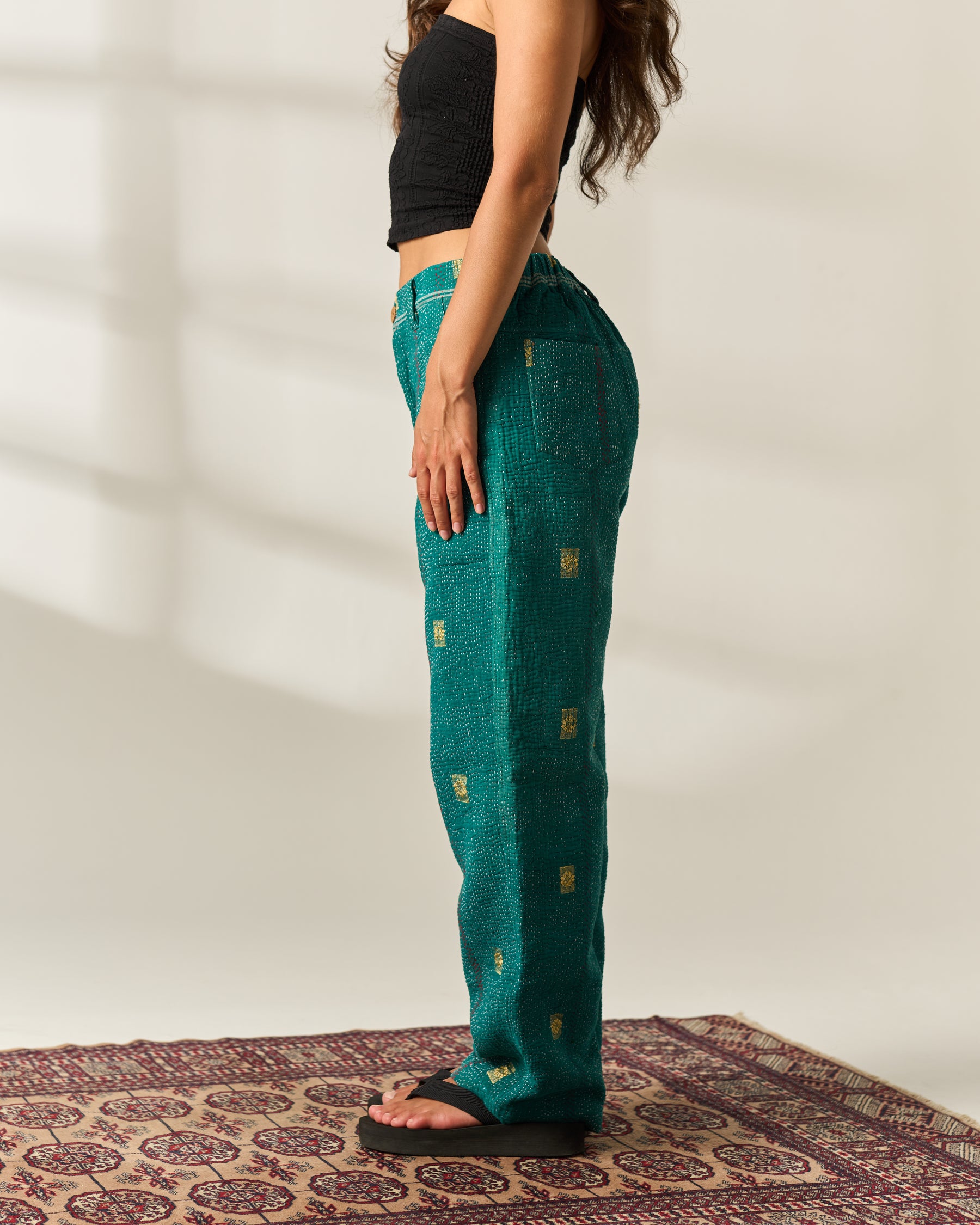 A woman stands in profile wearing teal-green wide-leg vintage Kantha pants with small gold rectangular hand-woven motifs, paired with a black crop top and black sandals, standing on a patterned rug against a light beige wall.