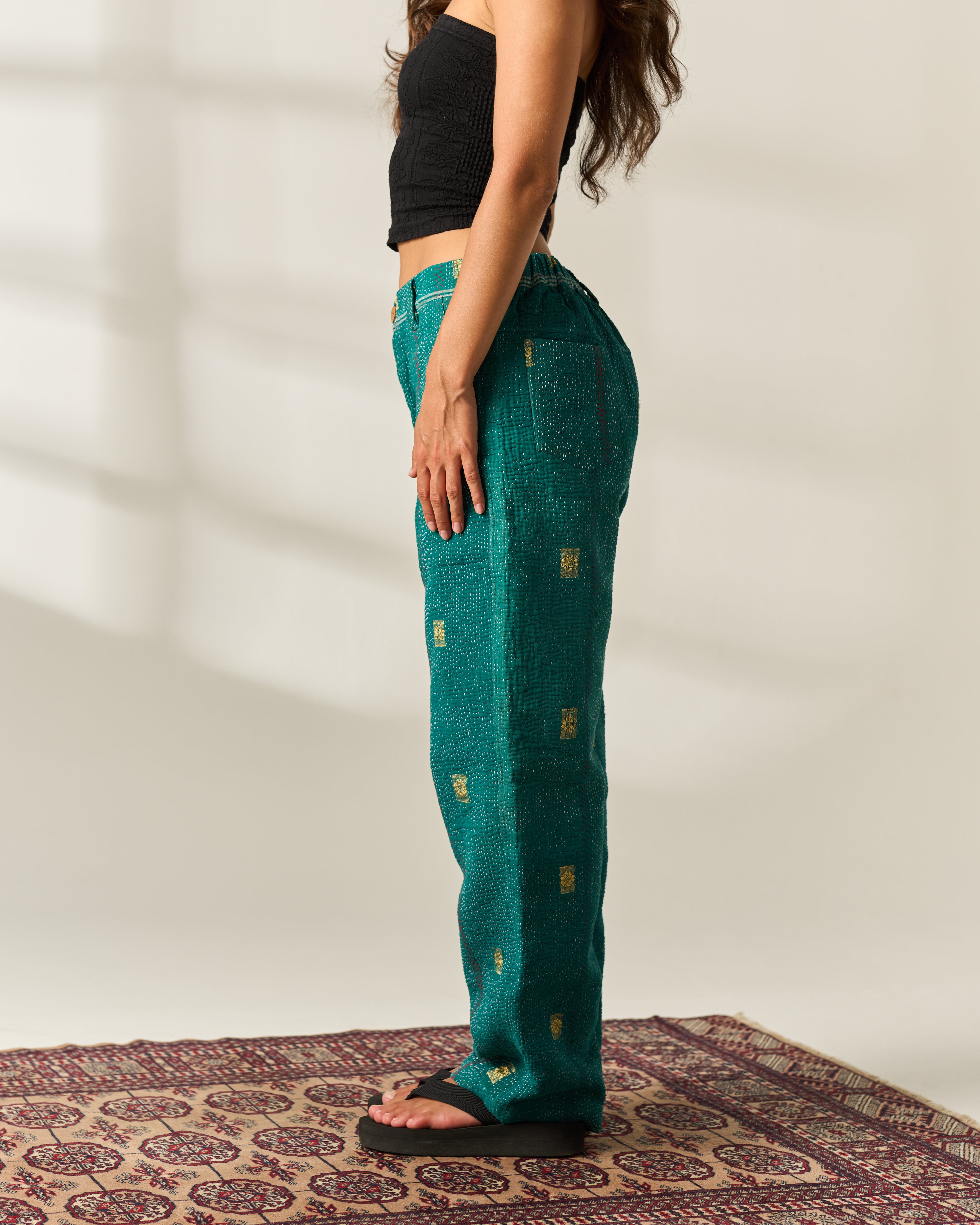 A woman stands in profile wearing teal-green wide-leg vintage Kantha pants with small gold rectangular hand-woven motifs, paired with a black crop top and black sandals, standing on a patterned rug against a light beige wall.