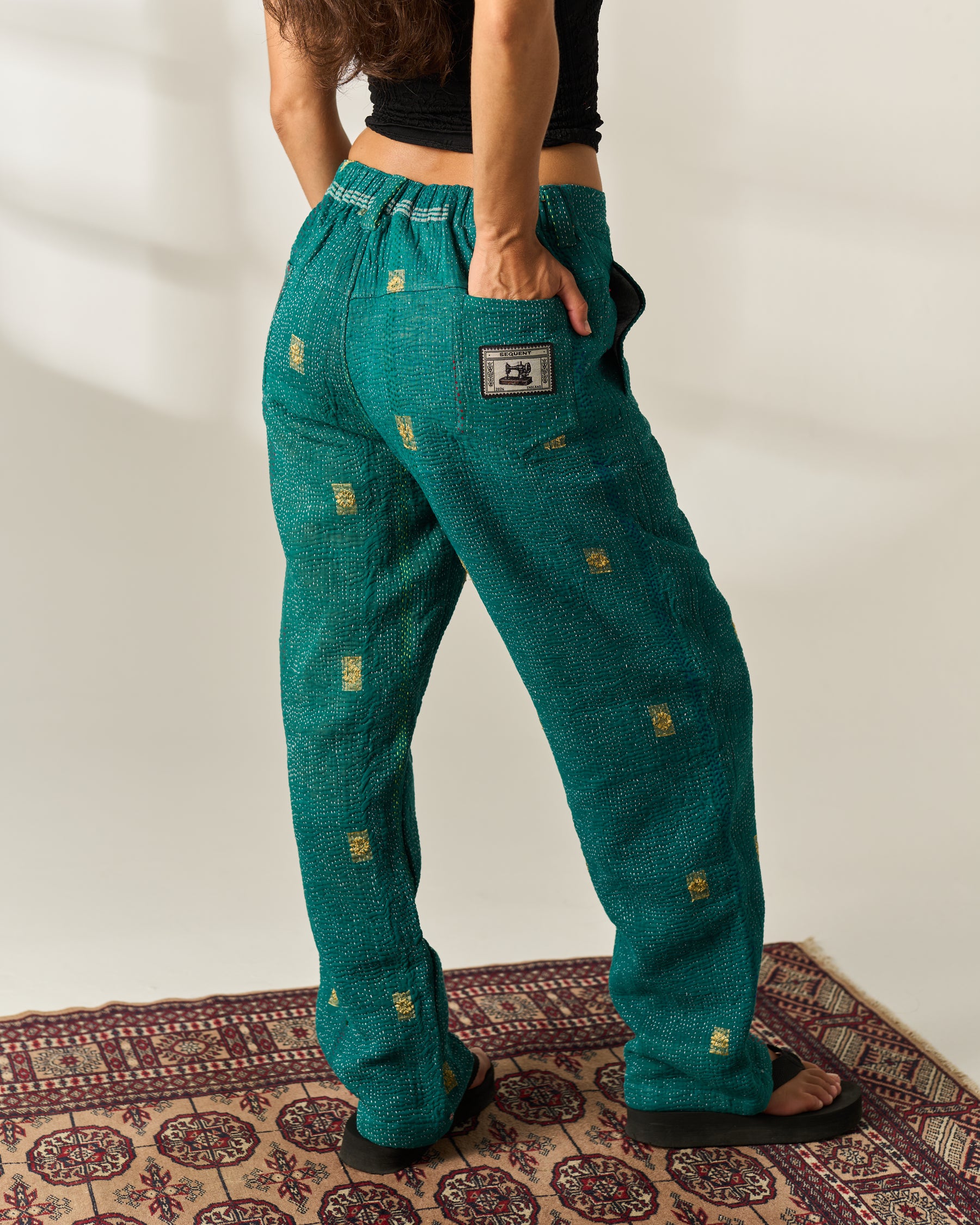 A person wearing teal green vintage Kantha pants with small gold geometric prints, shown from the back in a studio setting with a patterned rug on the floor.