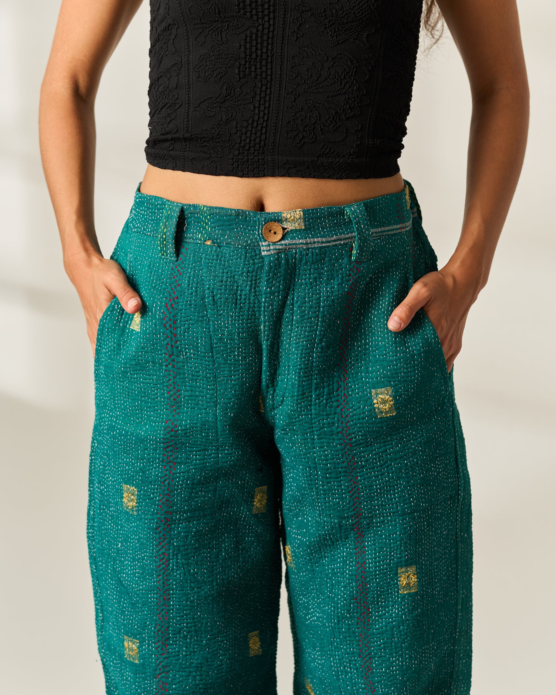A person wearing teal-green textured wide-leg vintage Kantha pants with small gold square hand-stitched motifs and a black cropped top, standing against a plain light background.