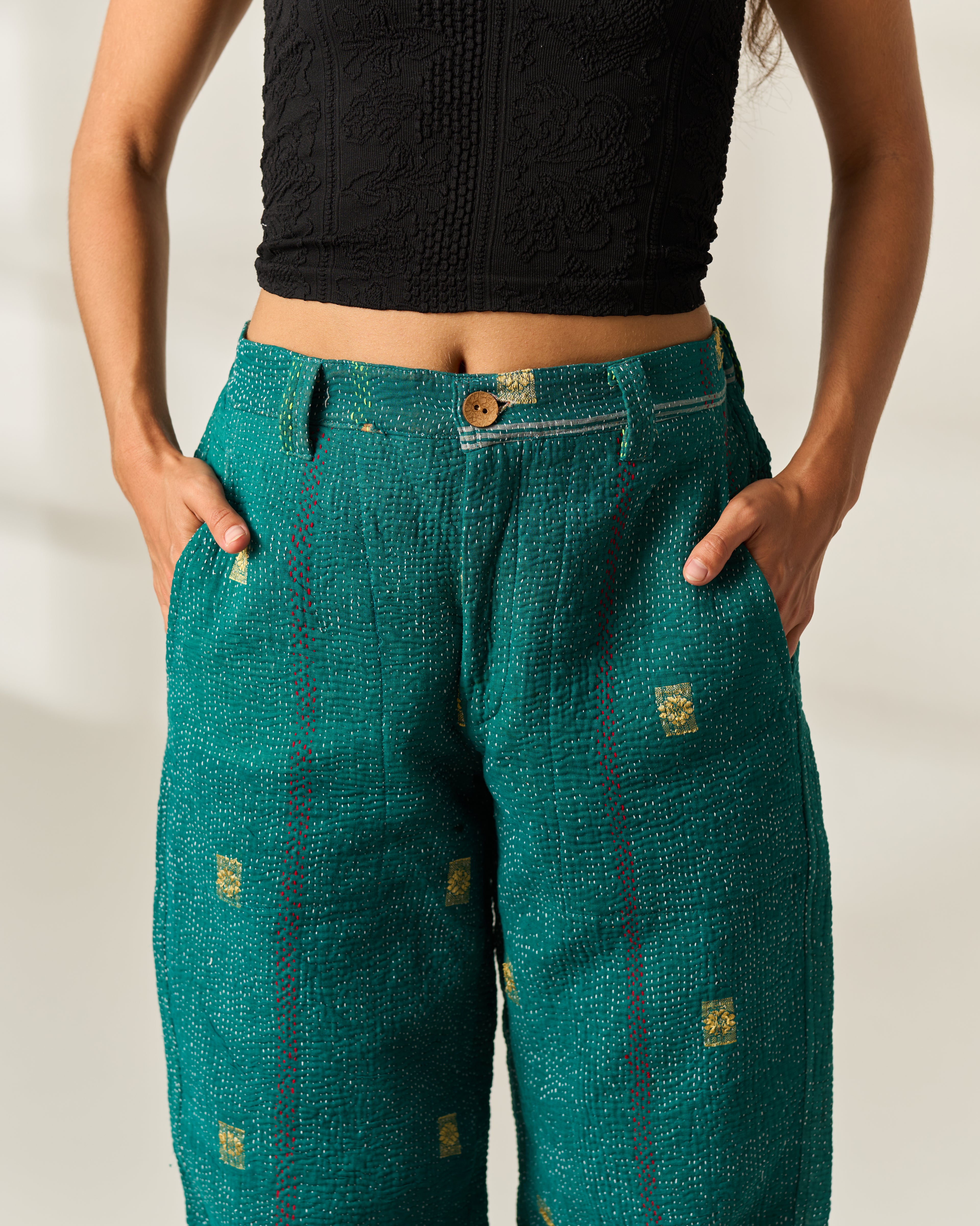 A person wearing teal-green textured wide-leg vintage Kantha pants with small gold square hand-stitched motifs and a black cropped top, standing against a plain light background.