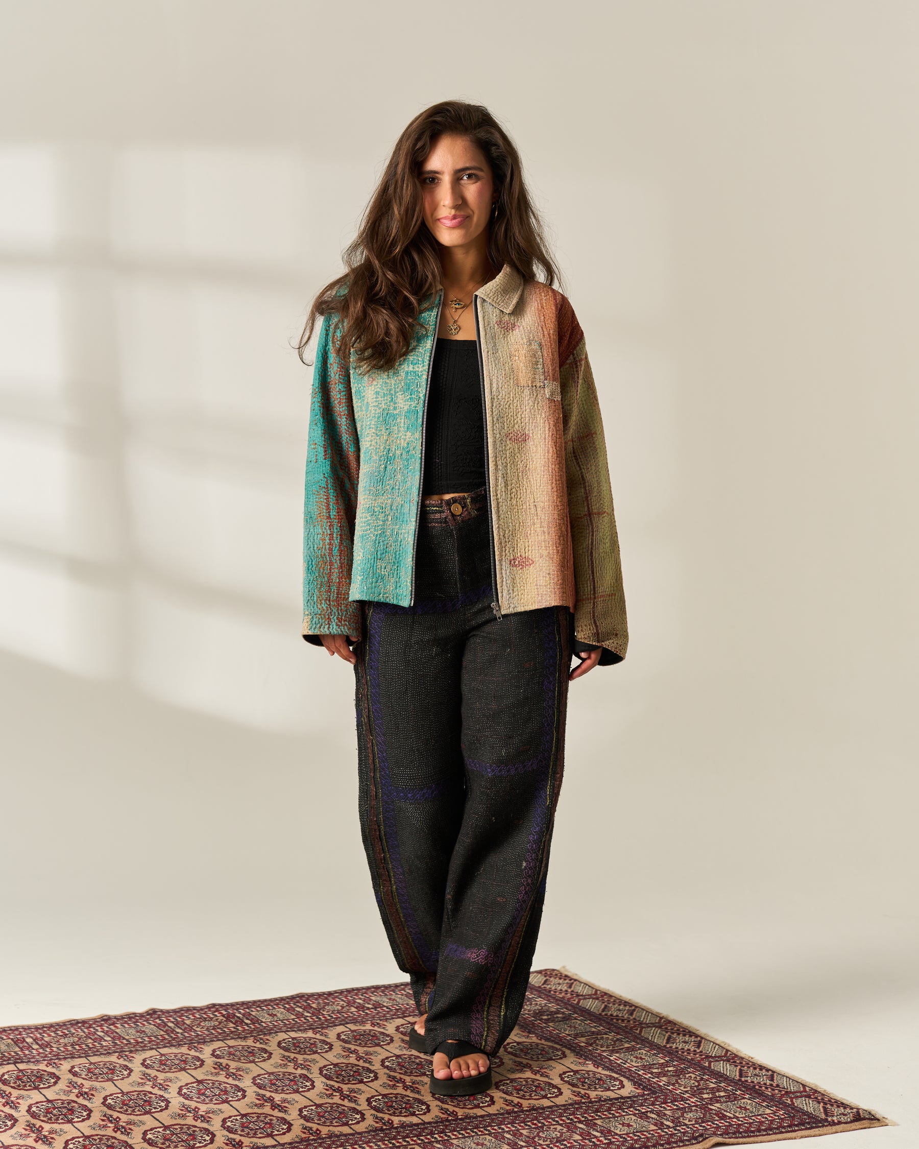A woman stands on a patterned rug in a studio setting, wearing a multi-color patchwork, upcycled Vintage Kantha jacket, with a zipped front, with turquoise, beige, and brown tones over a black top and dark wide-leg trousers.
