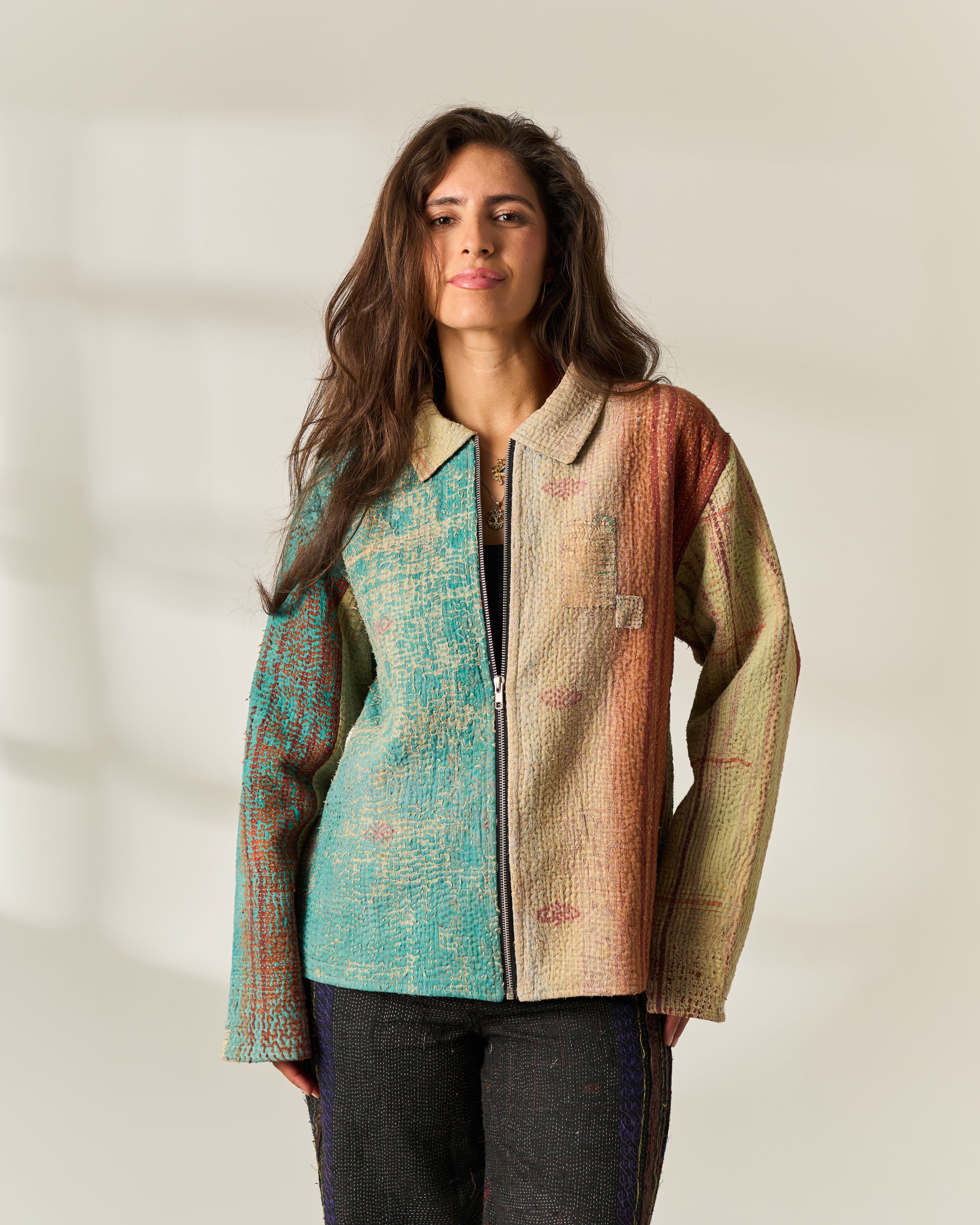 A woman modeling a zip-front upcycled Vintage Kantha jacket with a split color-blocked design: turquoise blue textured fabric on the left side and beige/rust textured fabric on the right side, paired with dark Vintage Kantha pants against a plain light beige studio background.
