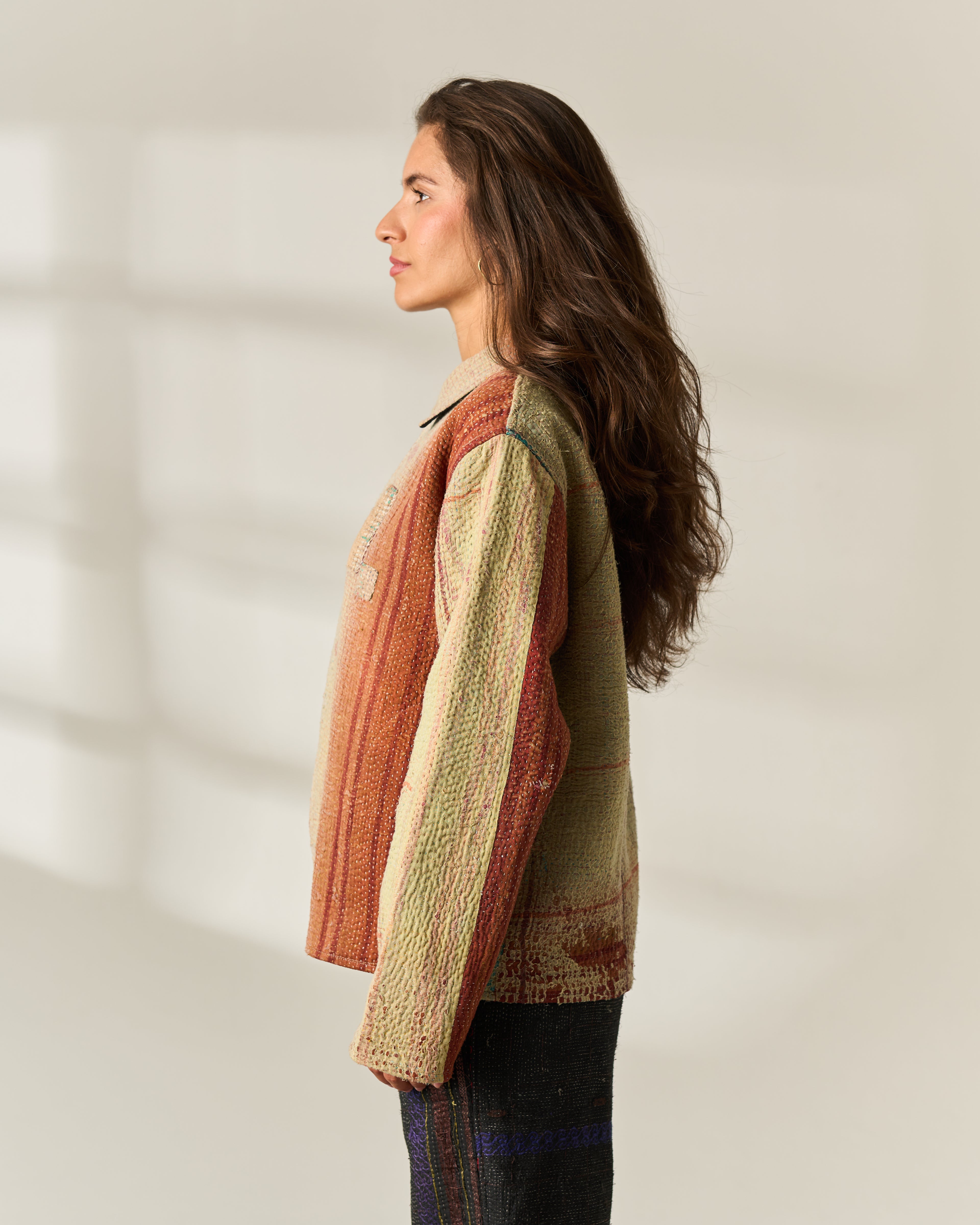 A woman in profile wearing a textured, multi-colored knit upcycled Vintage Kantha jacket with a mix of rust orange, beige, and olive green tones, paired with dark jKantha Trousers against a warm off-white background.