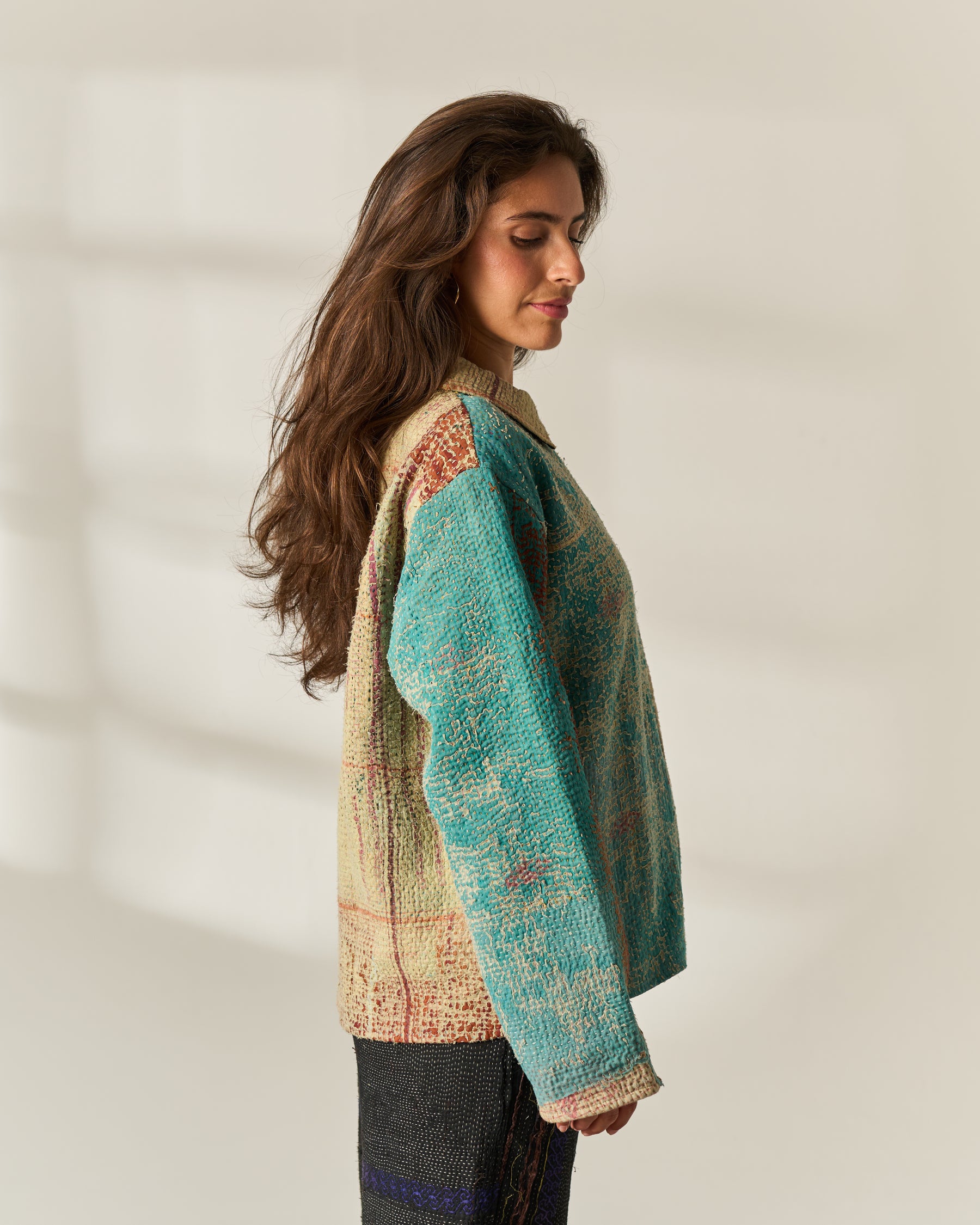 A woman in profile wearing a textured, multicolor upcycled Vintage Kantha jacket with a teal and beige pattern, standing against a warm off-white wall with soft window shadows.