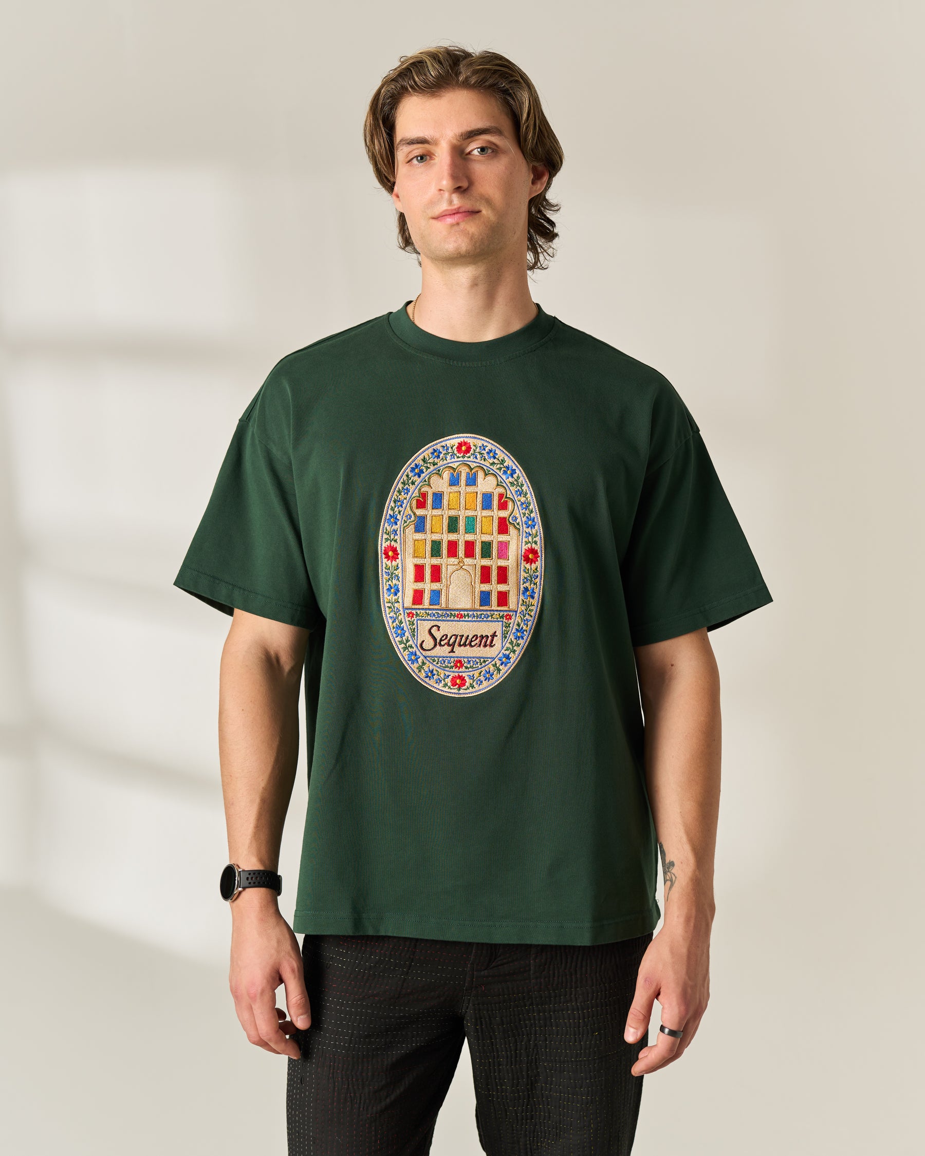 A man wearing a dark green crewneck Oversized 280 GSM t-shirt with a colorful circular graphic on the chest that reads 'Sequent' and features the windows of Jaipur. The background is a warm beige wall with soft window shadows.