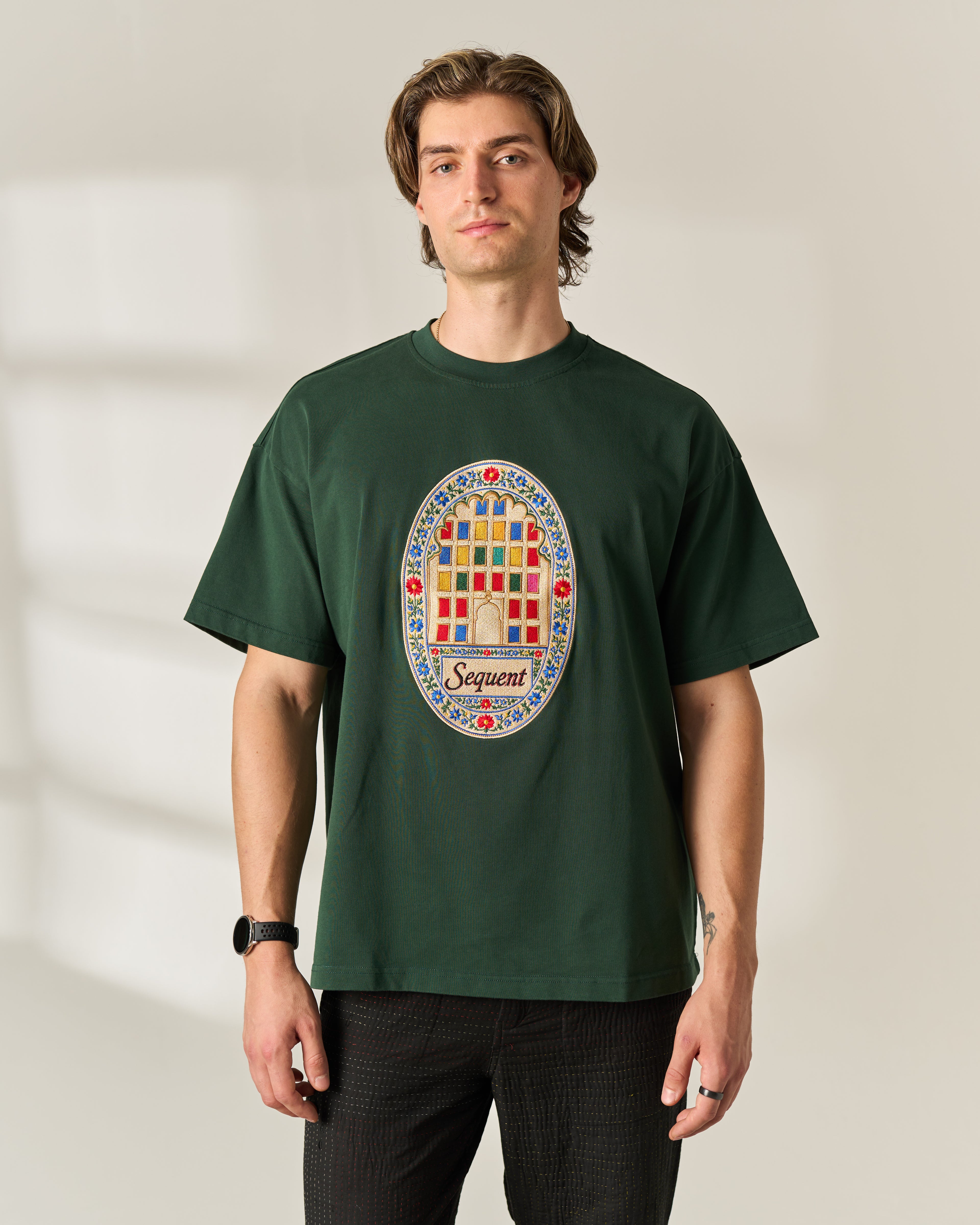 A man wearing a dark green crewneck Oversized 280 GSM t-shirt with a colorful circular graphic on the chest that reads 'Sequent' and features the windows of Jaipur. The background is a warm beige wall with soft window shadows.