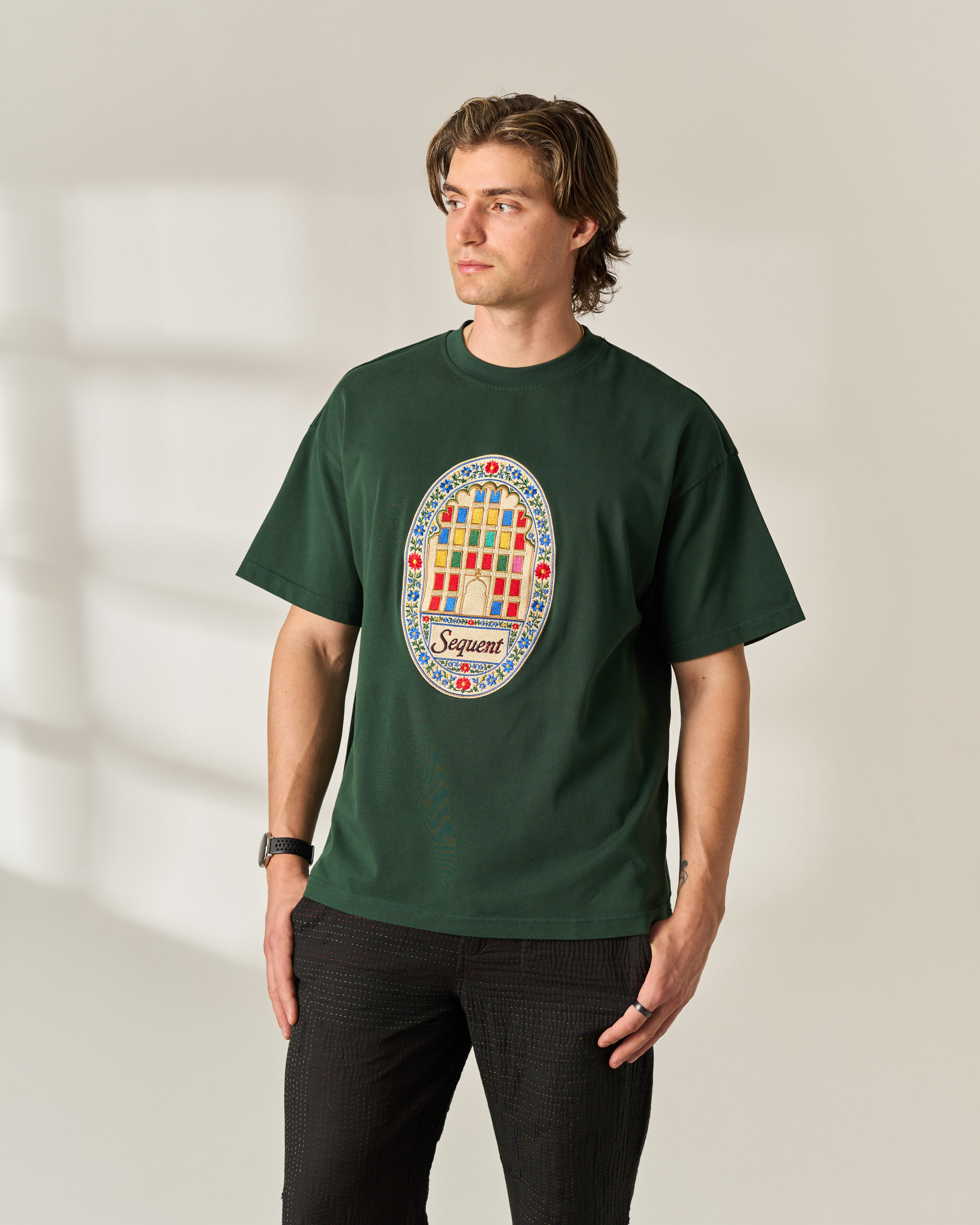 A man wearing a dark green crewneck Oversized 280 GSM Cotton t-shirt with a colorful circular graphic on the chest featuring the word 'Sequent' and a grid pattern, paired with black pants, standing against a warm beige background with soft window shadows.