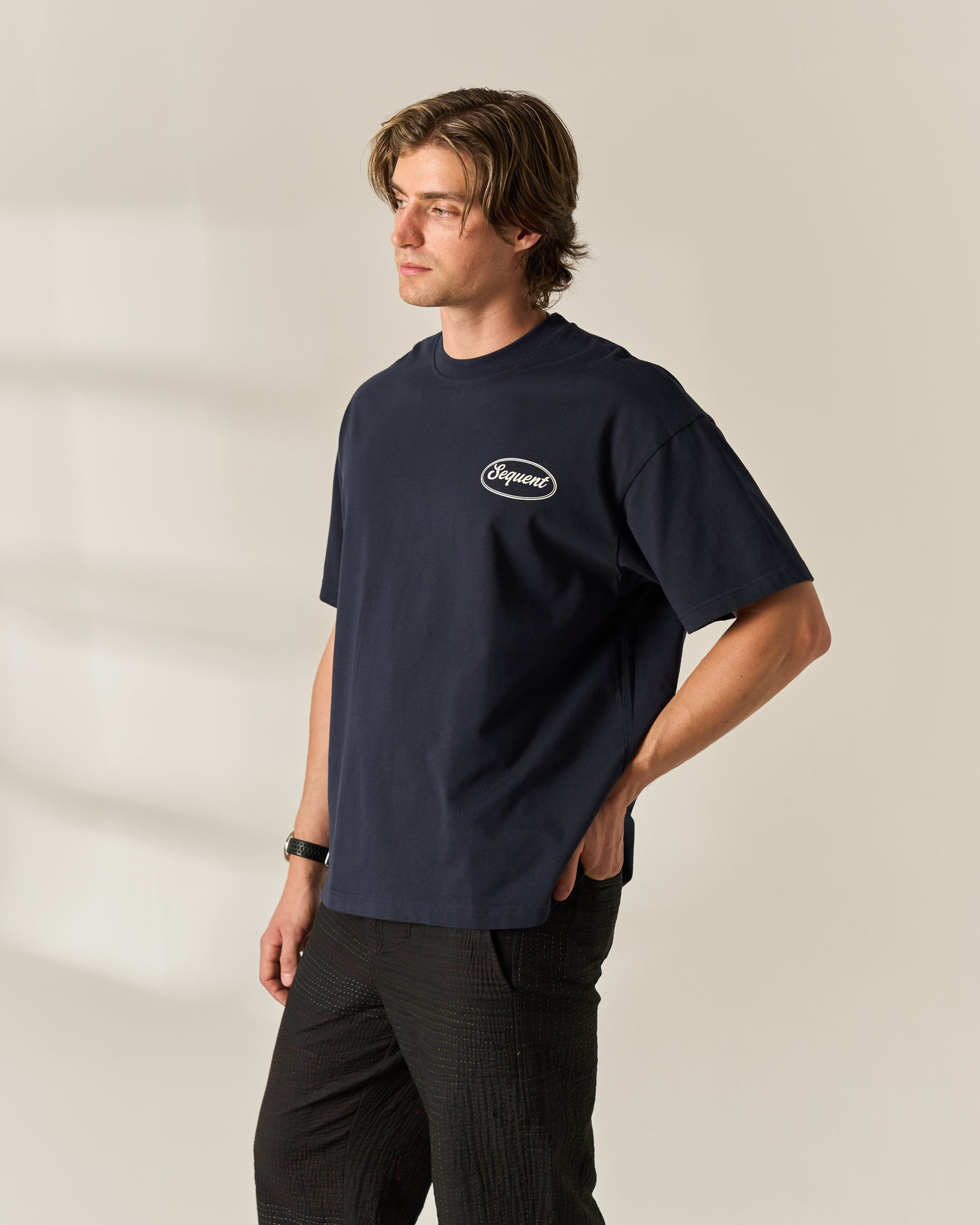 A man wearing a navy blue crewneck t-shirt with a small white oval 'Sequent' logo on the left chest, paired with dark gray pants, standing against a warm beige studio background.