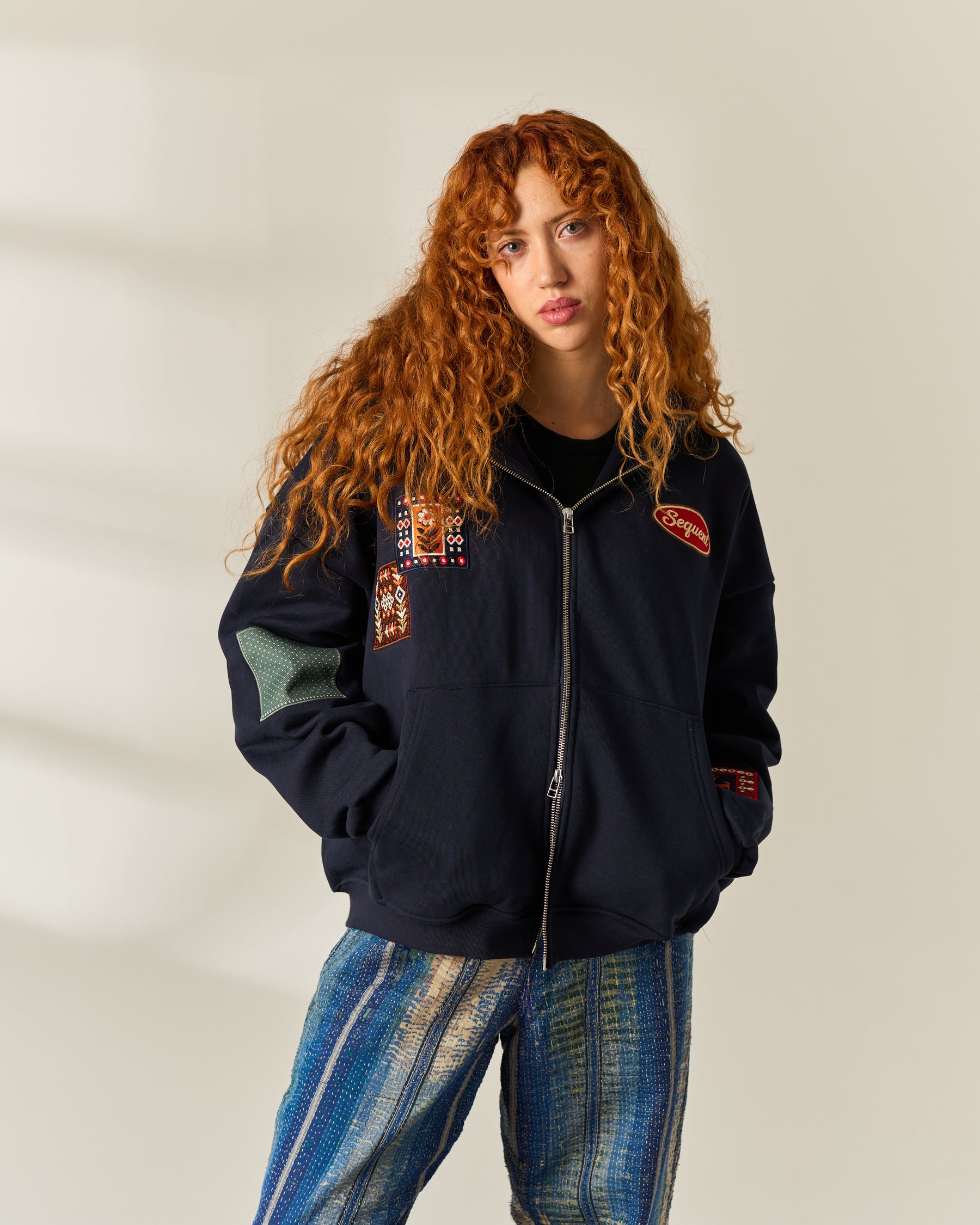 A model with long, curly red hair wearing a navy blue zip-up jacket with colorful embroidered patches on the chest and sleeves, paired with blue and white striped pants, set against a warm beige background with soft shadows.